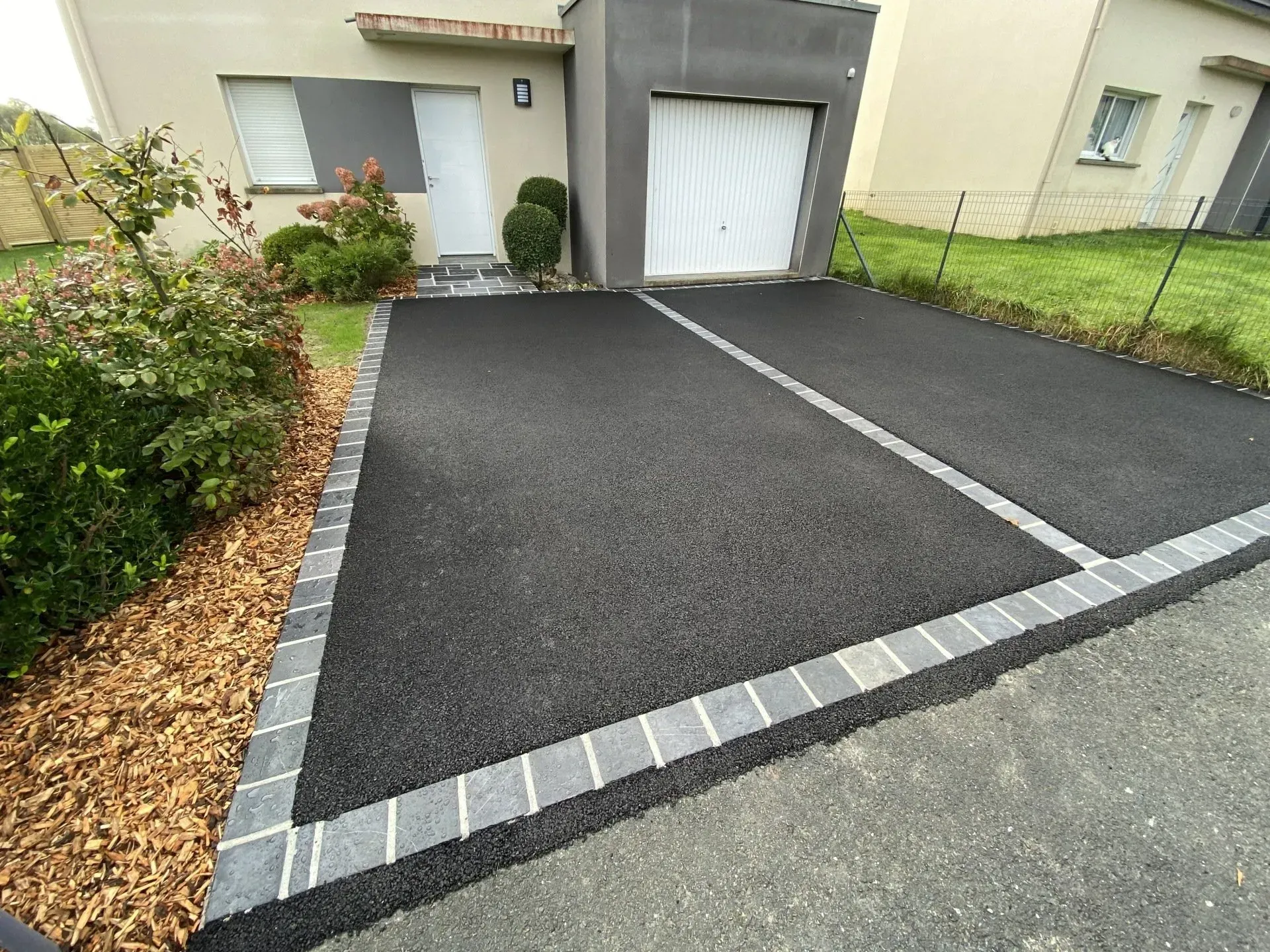 Allée carrossable noire fraîchement pavée avec bordures grises devant une maison et un garage.