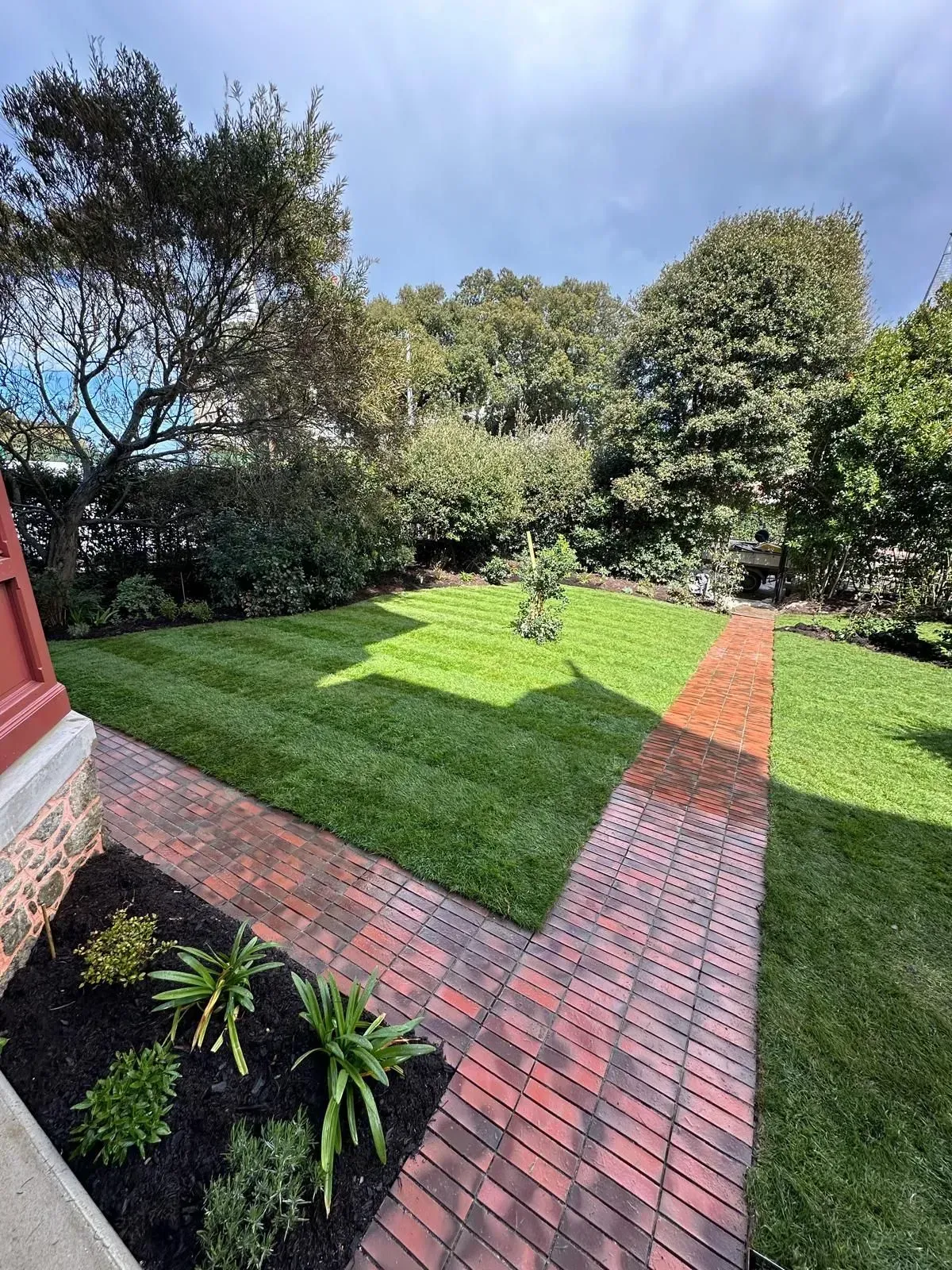 Allée de briques traversant un jardin verdoyant et ensoleillé, à côté d'une maison rouge et d'arbres.