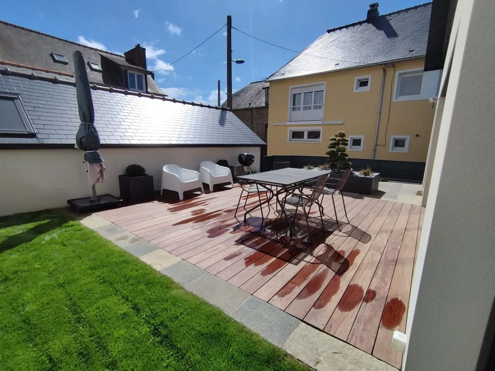 Terrasse avec table et chaises d'extérieur sur une terrasse en bois bordant une pelouse verte, sous un ciel d'un bleu éclatant.