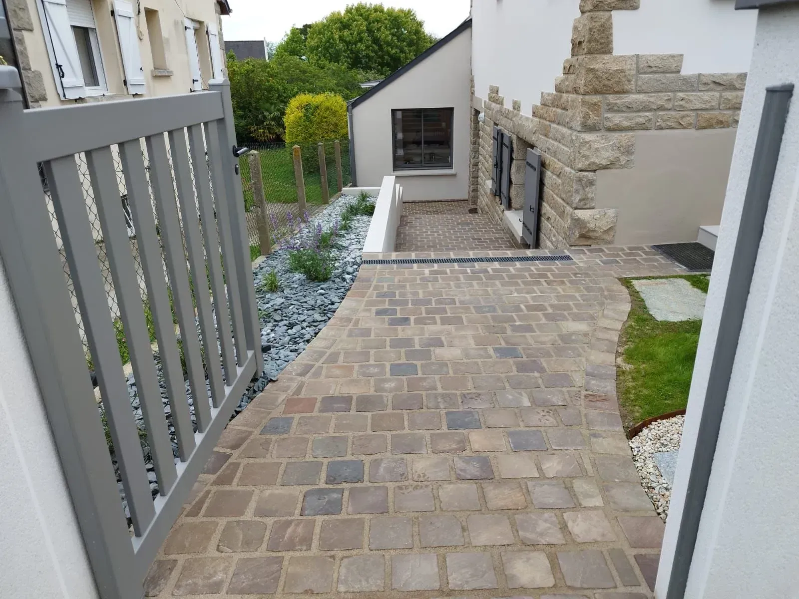 Étroit chemin pavé longeant une maison, avec une clôture grise et un mur de pierre.
