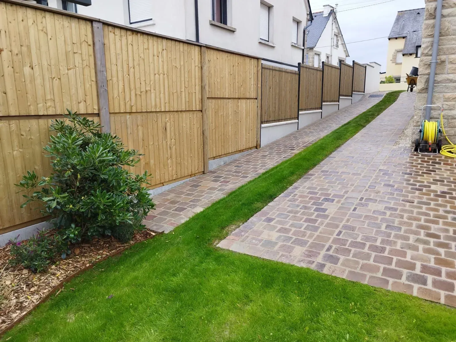 Ruelle résidentielle bordée de clôtures en bois, de bandes de gazon et d'un chemin pavé longeant les maisons.
