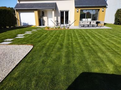 Jardin moderne avec pelouse verte à rayures, allée en pierre, chaises de patio et maison aux murs jaunes et blancs.