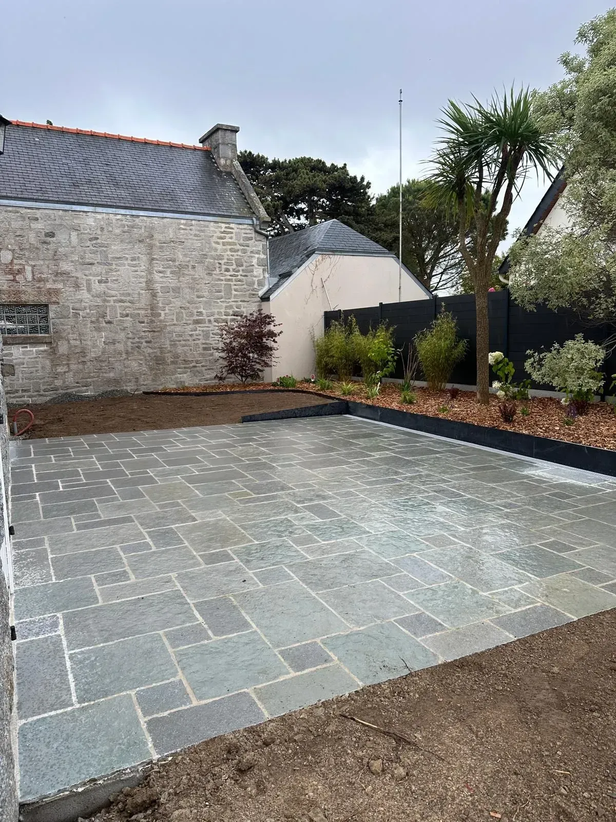 Terrasse en pierre mouillée jouxtant une maison en briques blanches, avec des parterres de jardin et des arbres après la pluie.