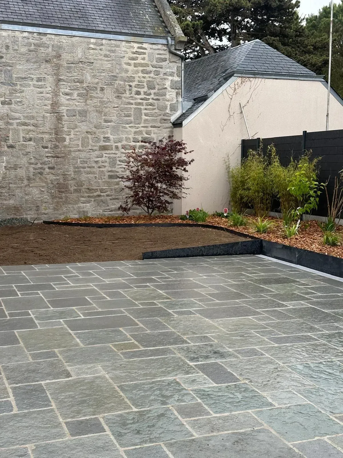 Cour en pierre avec pavage gris, parterres de fleurs surélevés et mur de briques jouxtant une maison.
