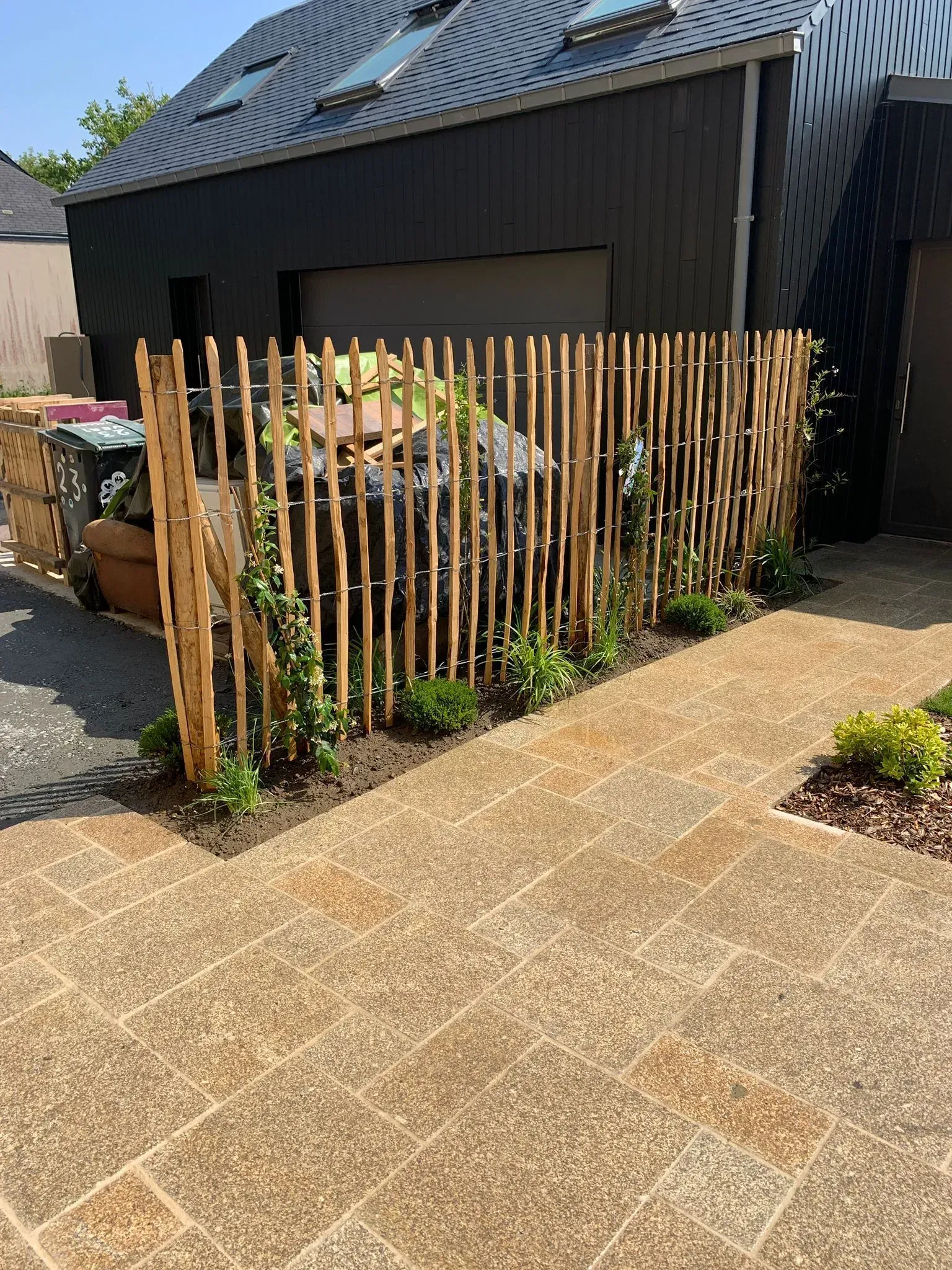 Clôture en bois entourant un jardin jouxtant une maison noire et une allée de gravier.