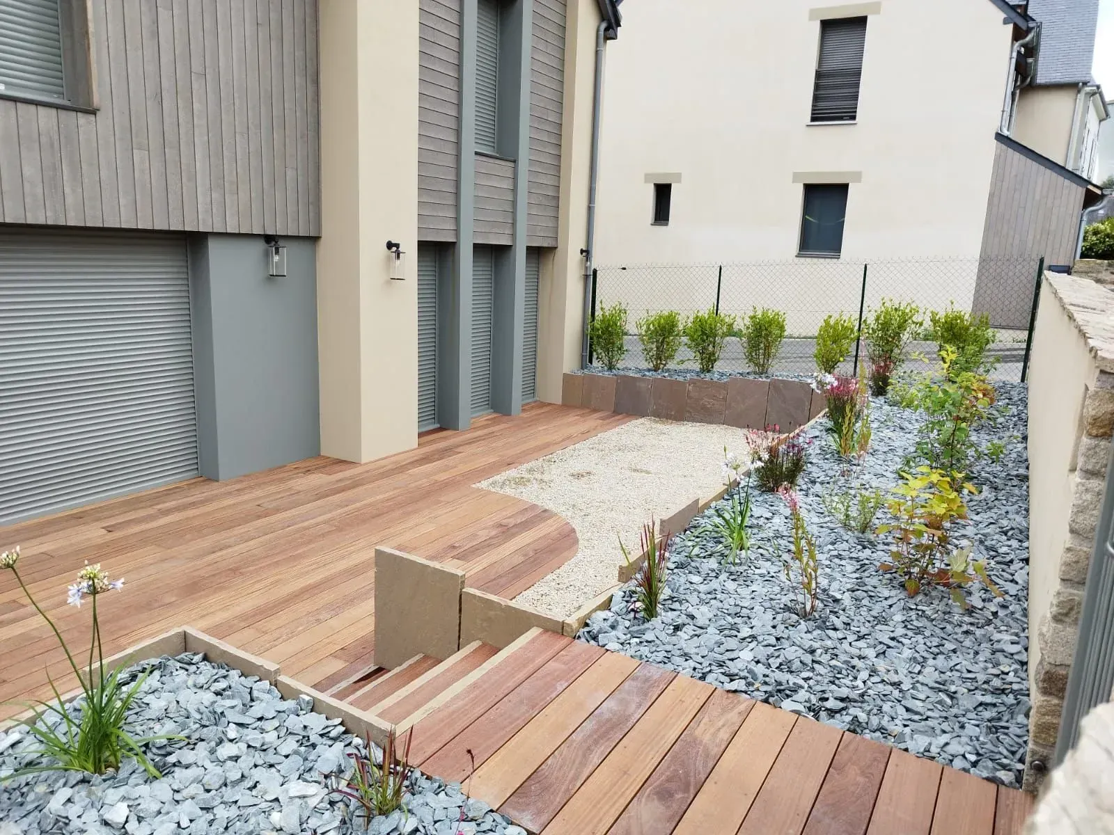 Patio moderne dans une cour arrière avec terrasse en bois, jardin de gravier et plantes en pot à côté de bâtiments beiges.