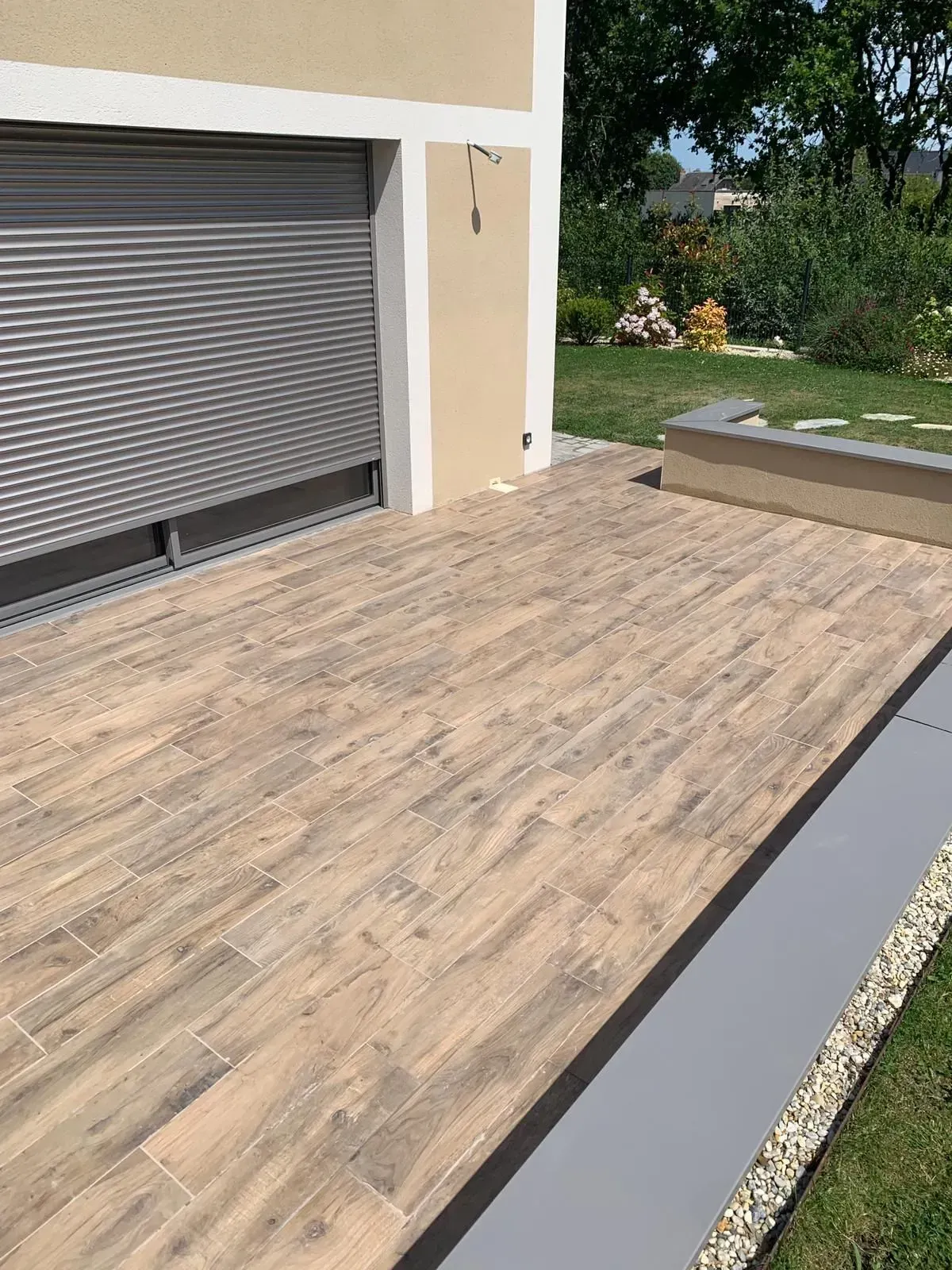Terrasse avec dalles imitation bois à côté d'une maison et d'une porte à volets gris fermée.