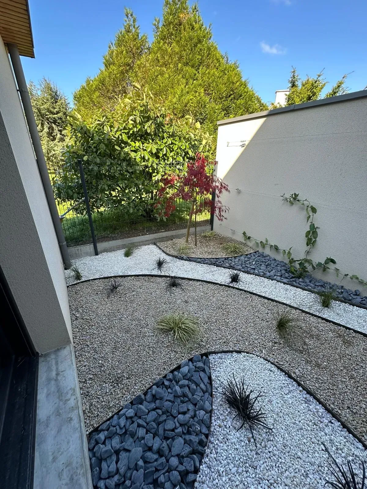 Jardin de gravier avec bordures courbes en pierre noire, arbustes et un mur blanc jouxtant une terrasse.
