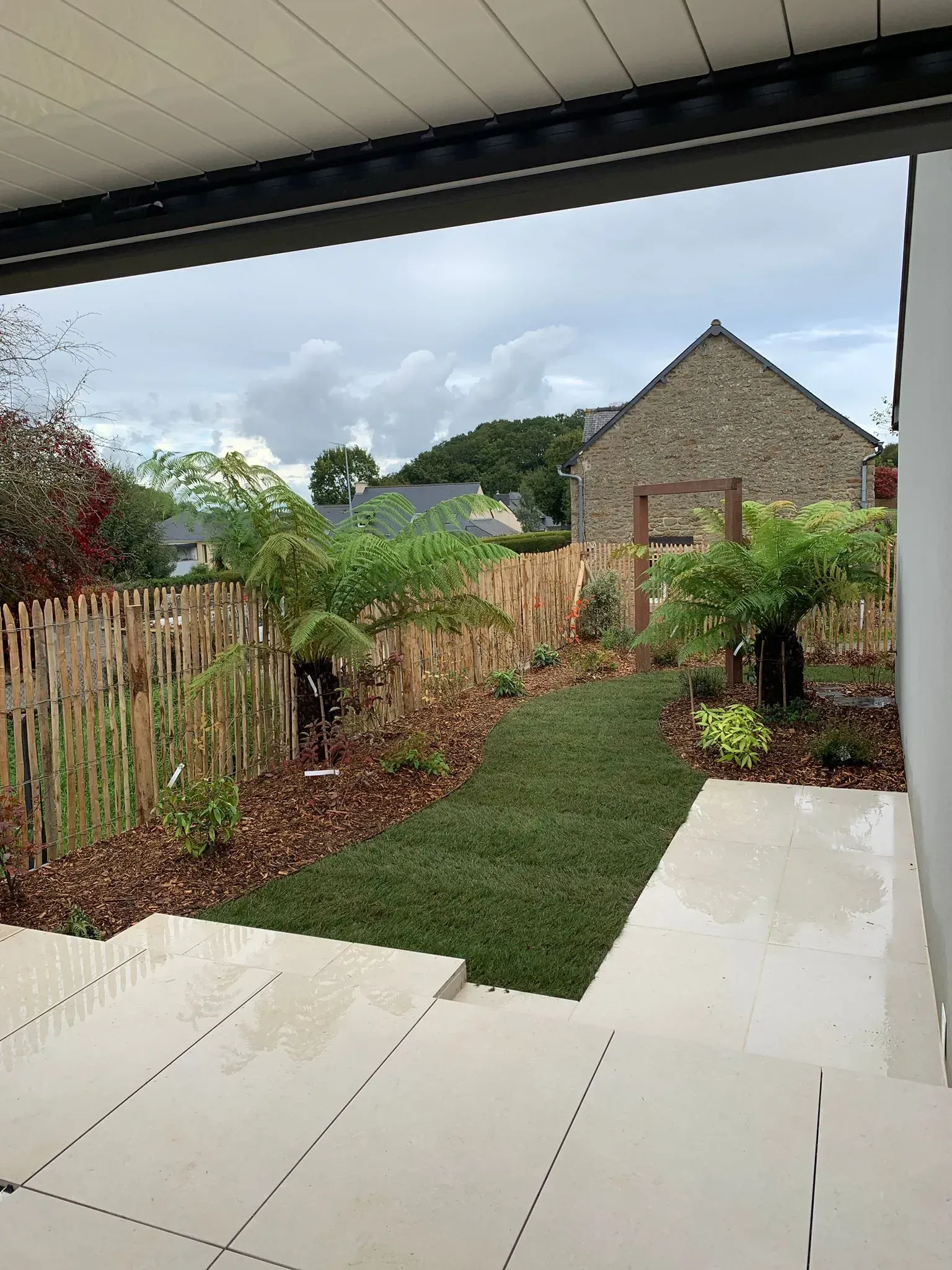Patio couvert donnant sur un étroit chemin de jardin, des haies et une maison en briques sous un ciel nuageux.
