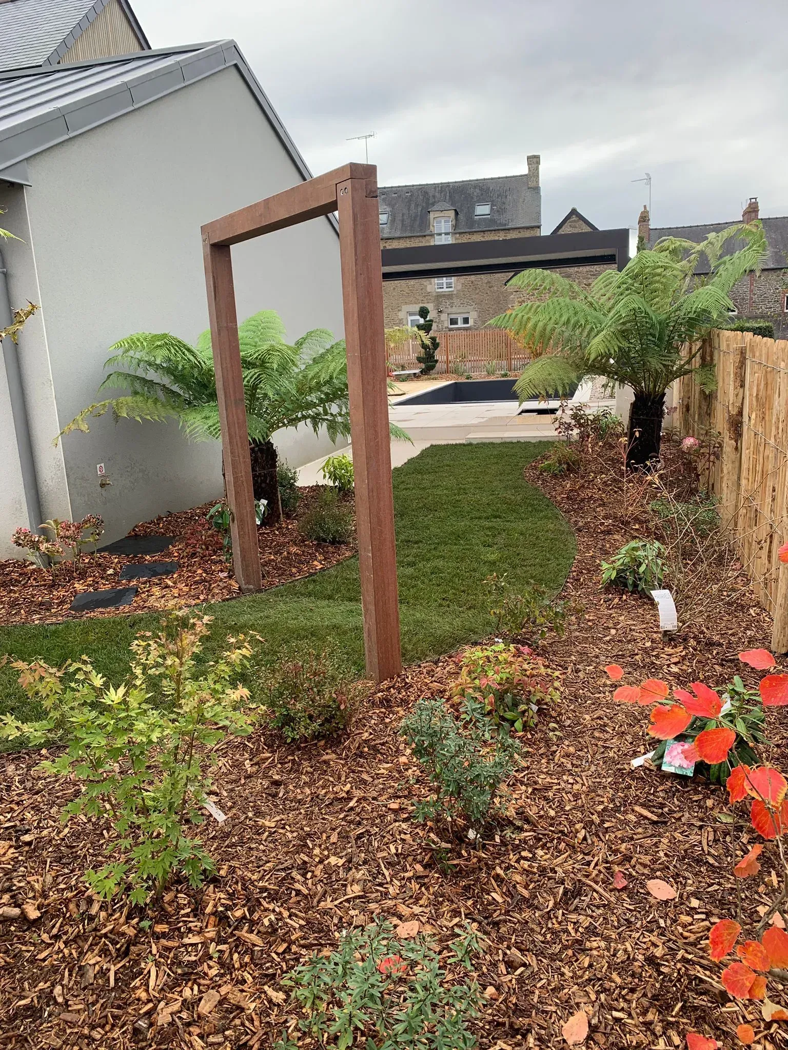 Allée de jardin avec une arche en bois, des arbustes verts et des fleurs rouges à côté d'une maison sous un ciel nuageux.