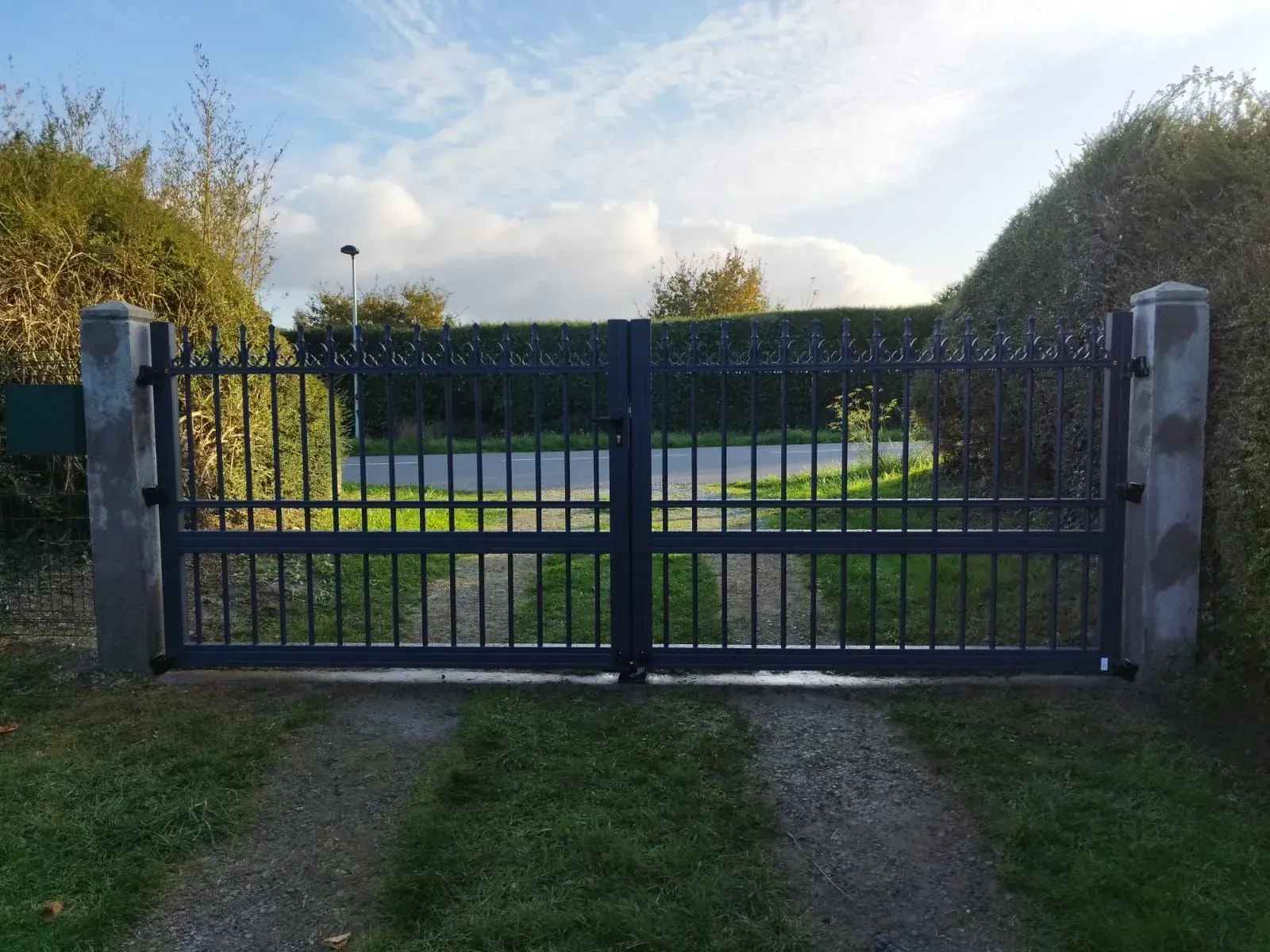 Un portail en métal noir barre une allée herbeuse, avec des poteaux en pierre et des arbres sous un ciel nuageux.