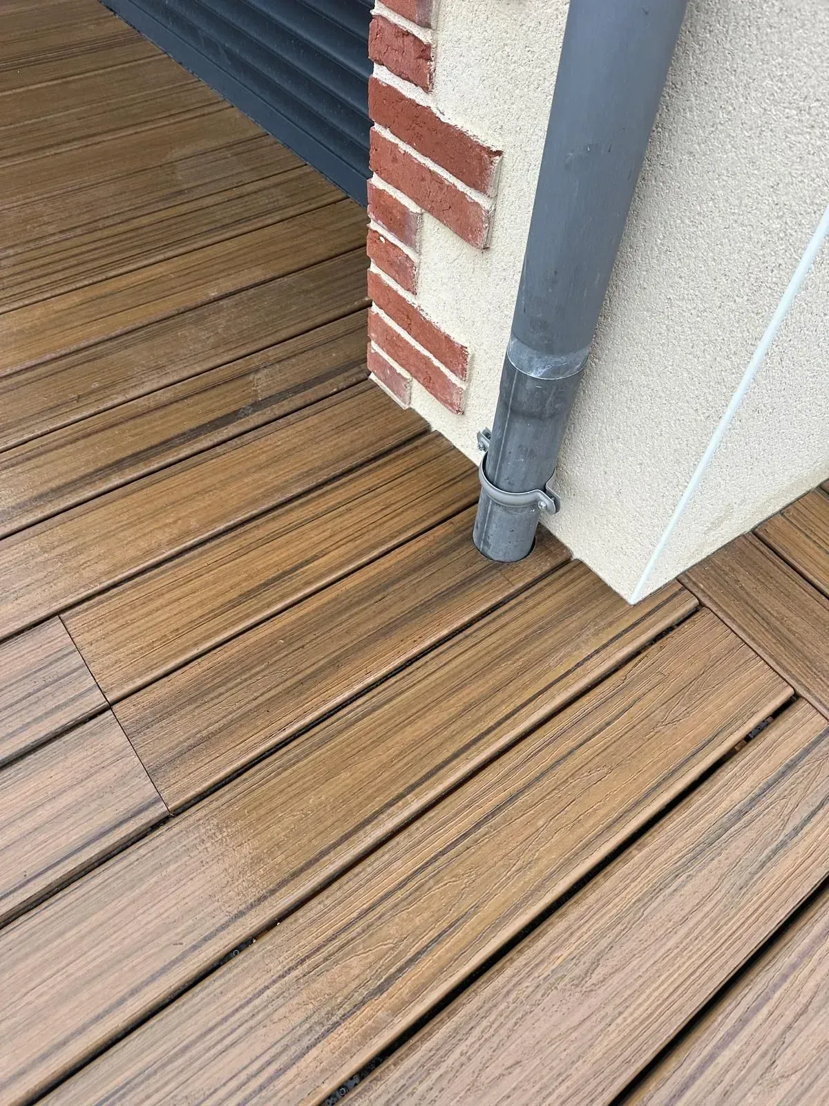 Des planches de terrasse en bois longent un mur de briques et un tuyau de descente gris à un angle de bâtiment.