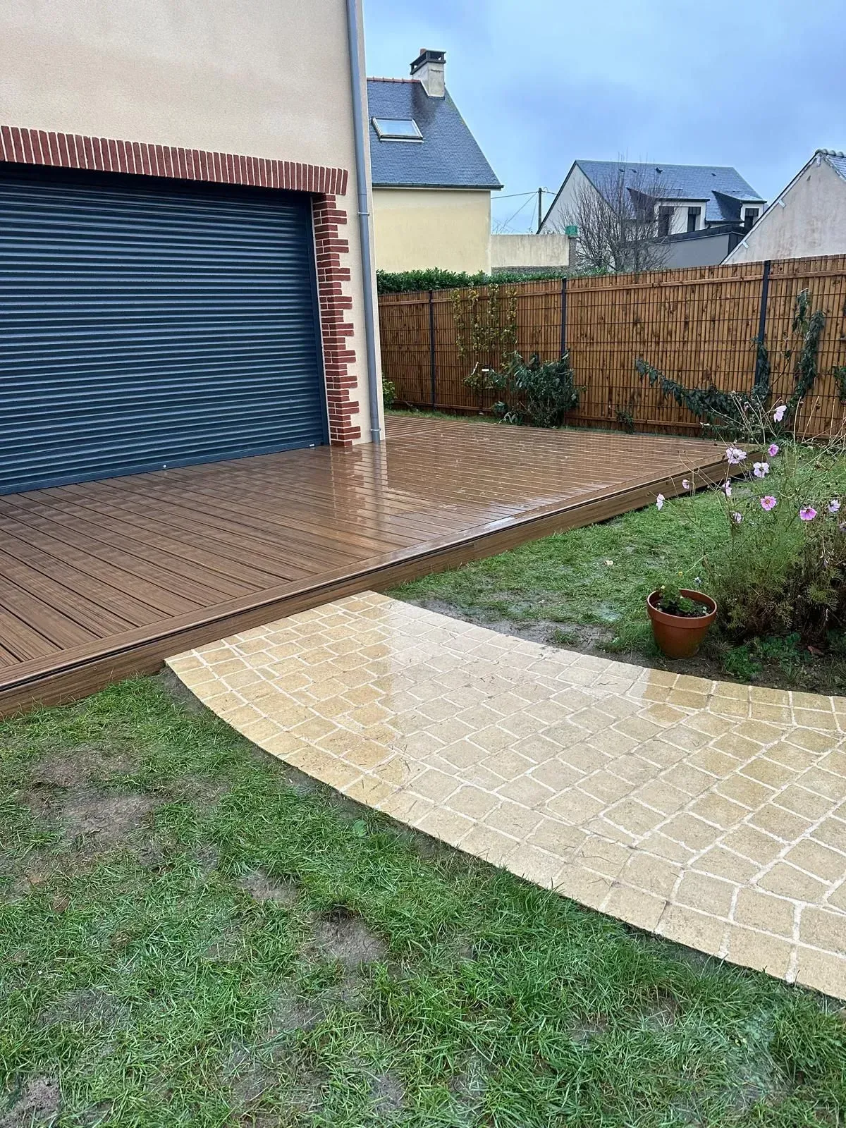 Terrasse arrière pluvieuse avec terrasse en bois mouillée, allée carrelée, porte de garage et petits parterres de jardin.