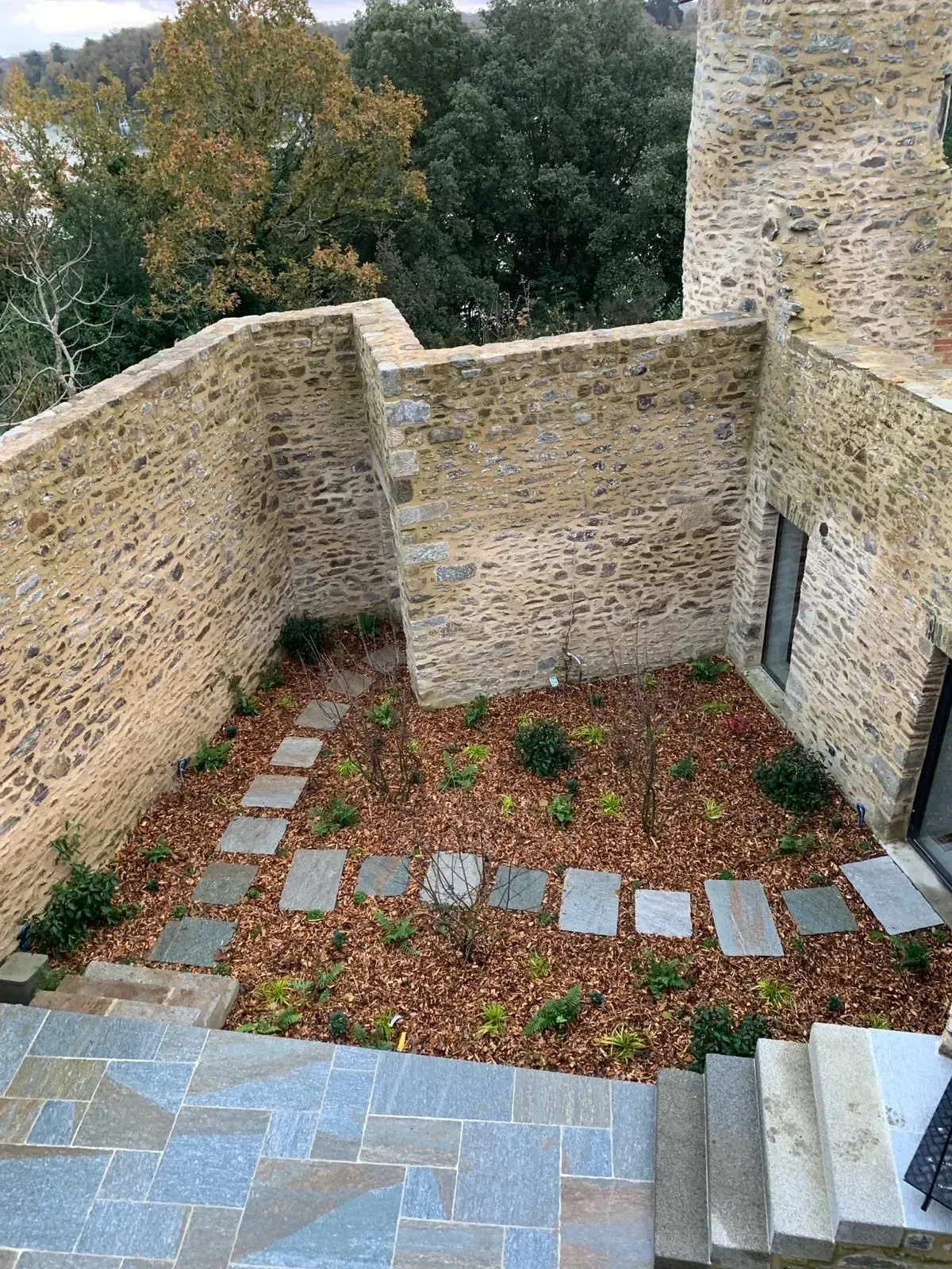 Cour en pierre avec des murets, des feuilles éparses et des marches descendant vers une zone pavée.