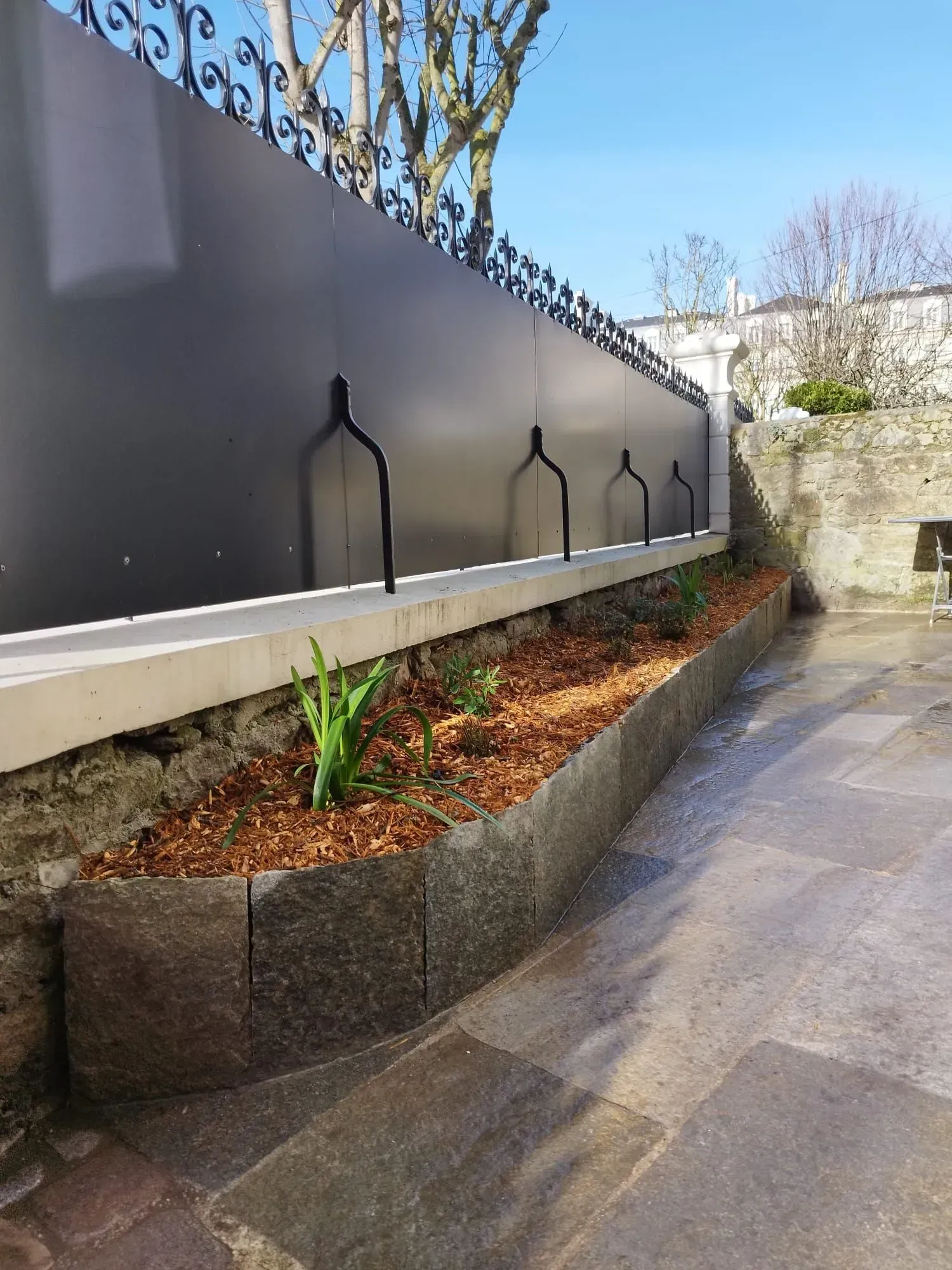 Allée paysagée moderne avec parterres surélevés, clôture sombre et pavage en pierre le long d'un mur.