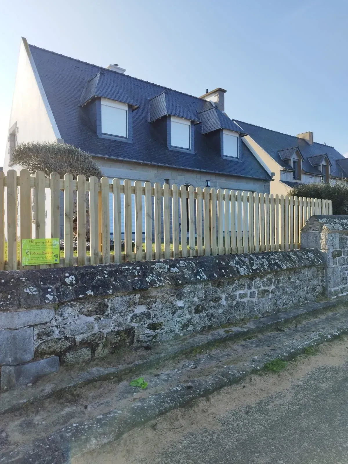 Maison en pierre au toit bleu derrière une clôture claire dans une rue ensoleillée.