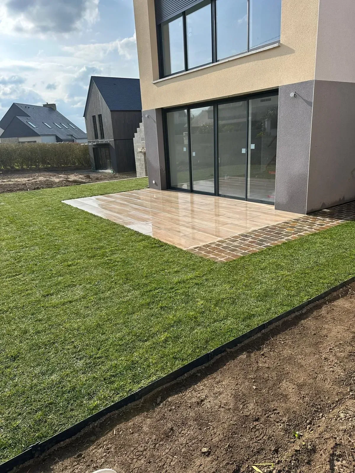 Extérieur de maison moderne avec de grandes portes vitrées, des dalles de terrasse et une pelouse verdoyante.