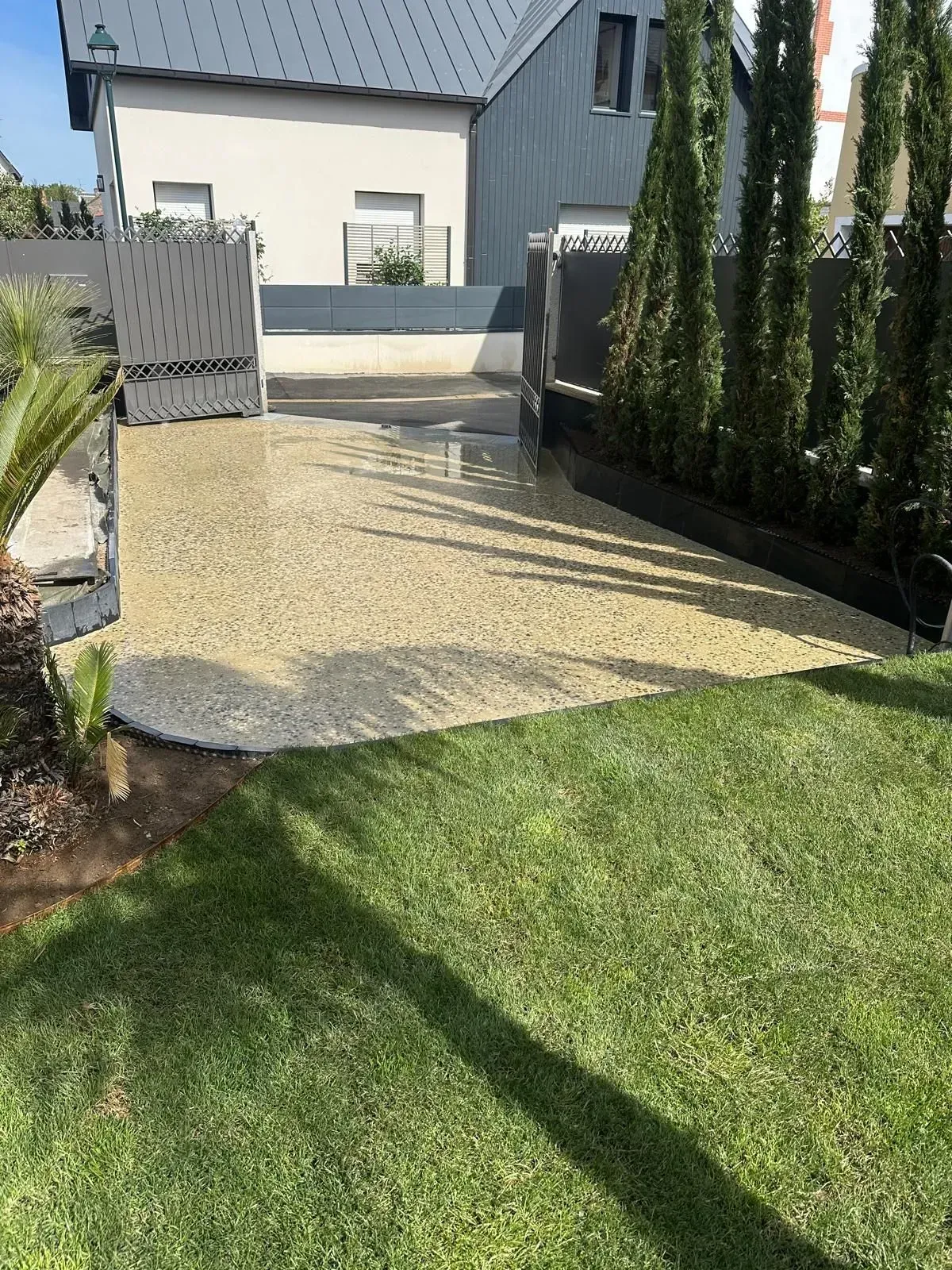 Petite terrasse en gravier bordant une pelouse verte, avec de grands cyprès et une maison moderne en arrière-plan.