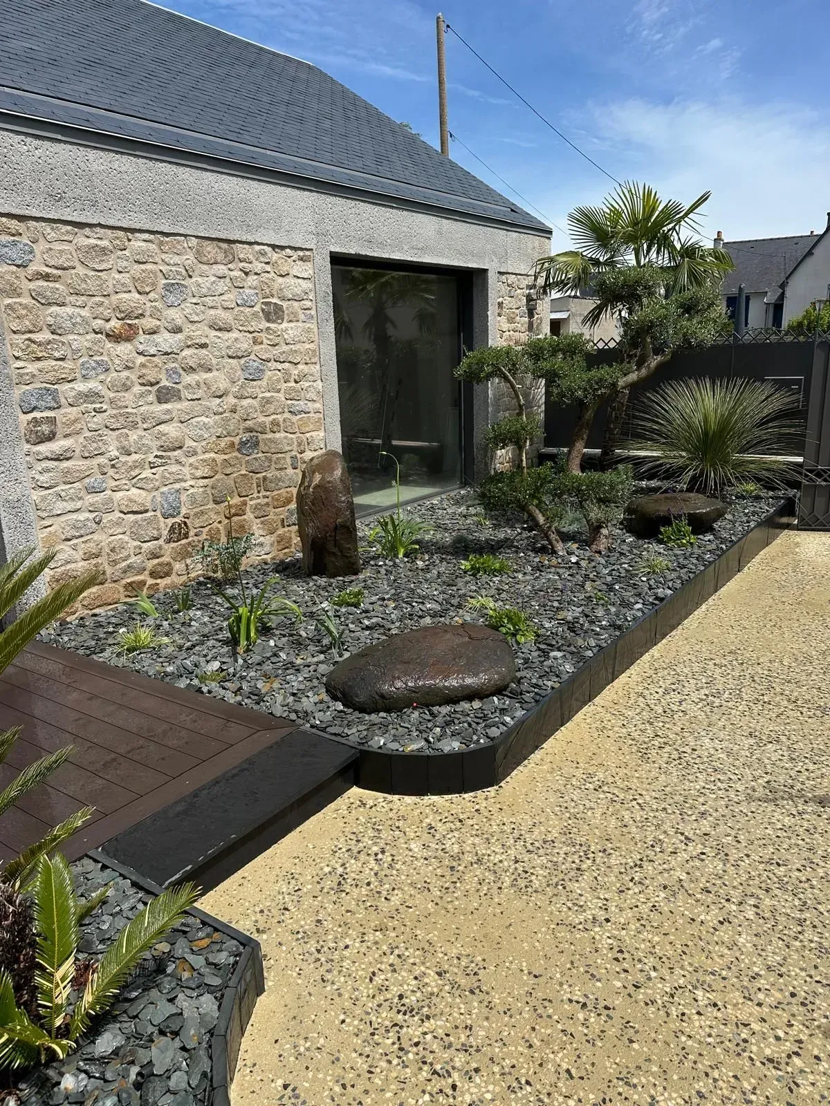 Maison en pierre avec un jardin de gravier, un petit étang et des plantes en pot au bord d'un chemin pavé.