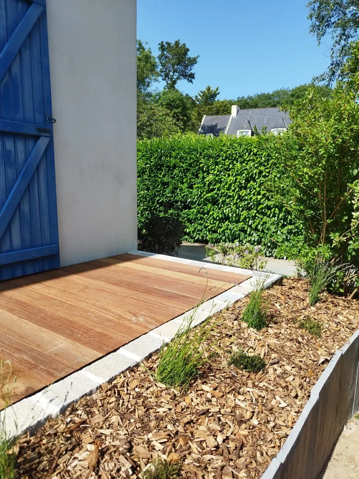 Porte bleue donnant sur une terrasse en bois, haie verte et parterre de jardin paillé baigné de soleil.