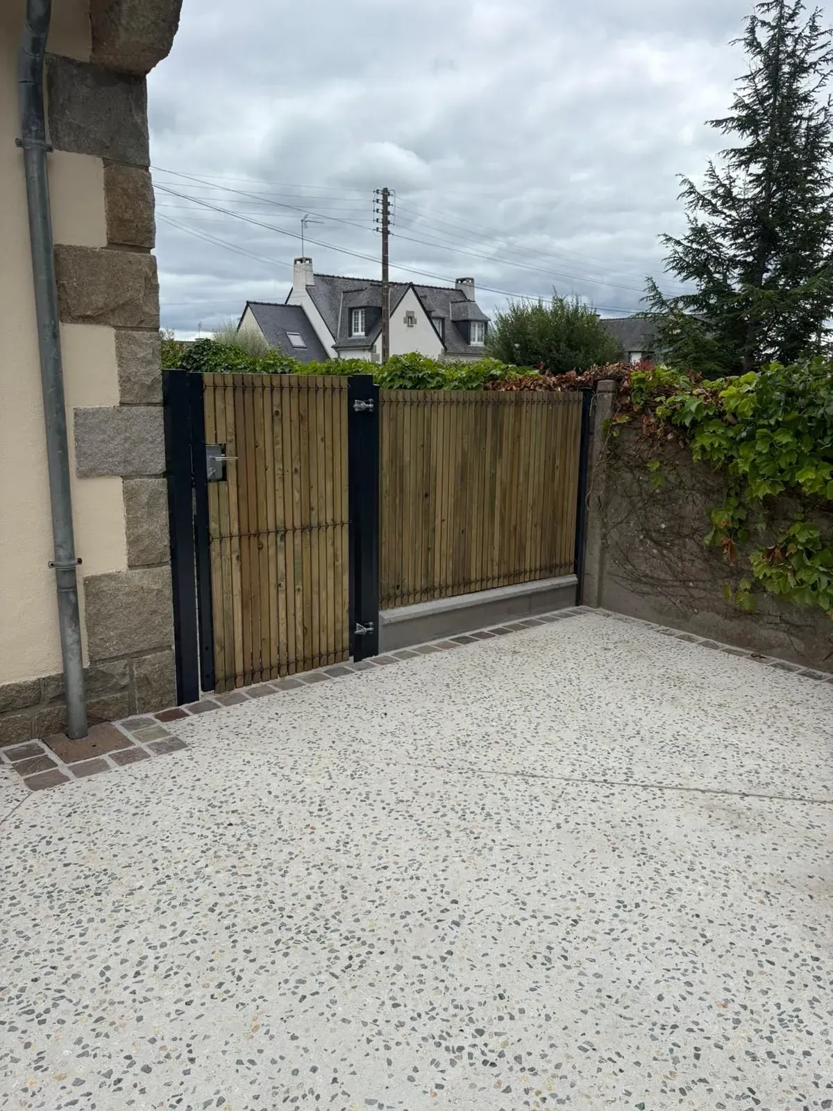 Portail en bambou jouxtant un mur de pierre, s'ouvrant sur une terrasse en béton moucheté sous un ciel nuageux.