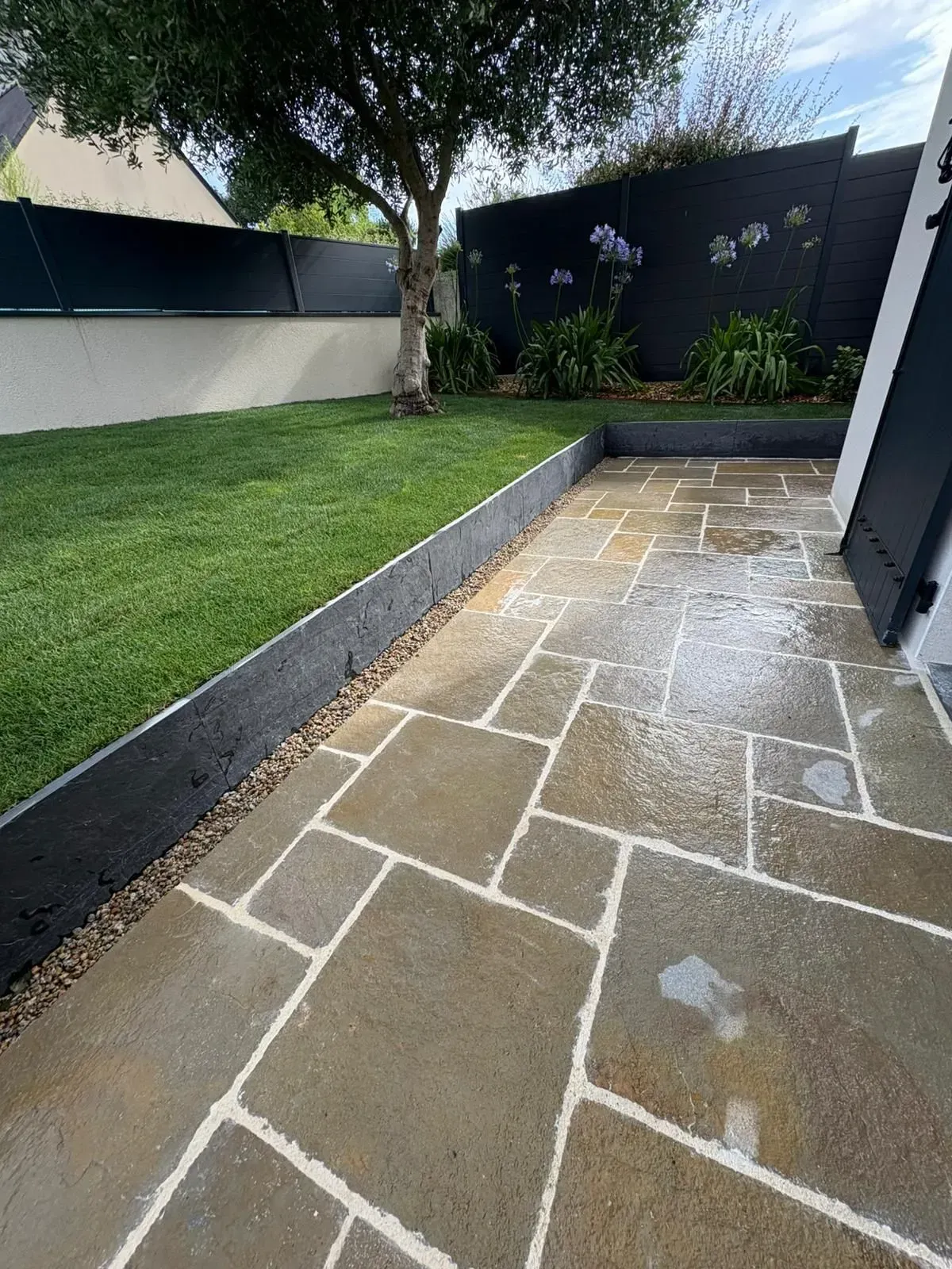 Jardin moderne avec pelouse verte, terrasse pavée, jardinière surélevée et clôture noire pour plus d'intimité.