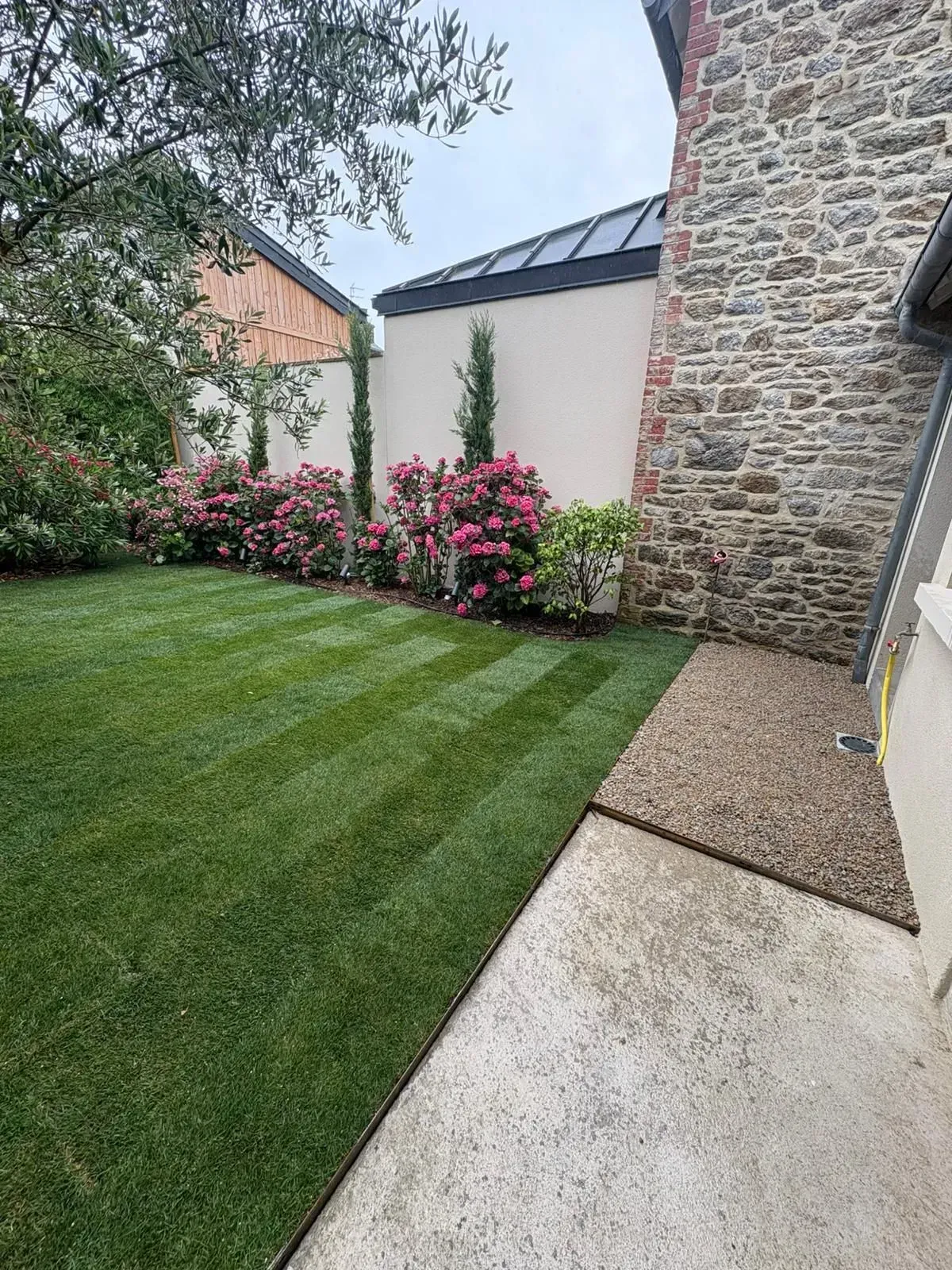 Jardin en pierre avec pelouse soignée, arbustes à fleurs roses et allée pavée.