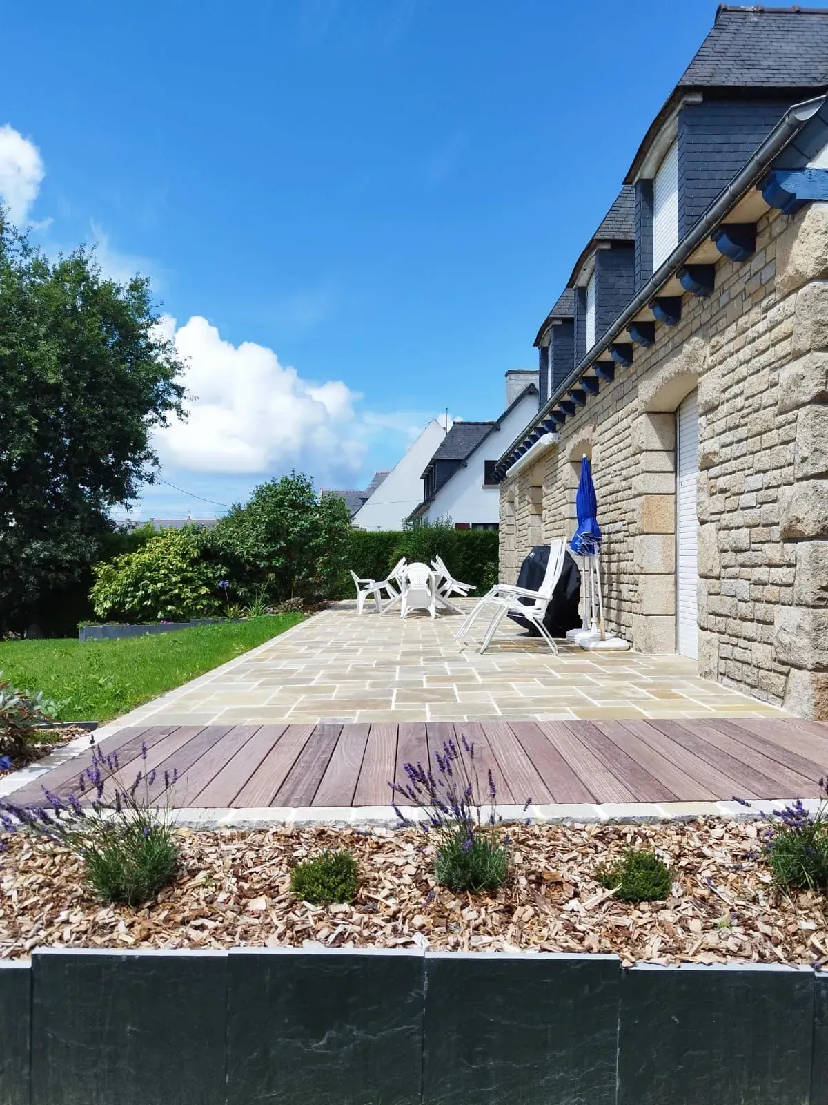 Patio en pierre avec chaises longues blanches, terrasse en bois et jardin sous un ciel d'un bleu éclatant.