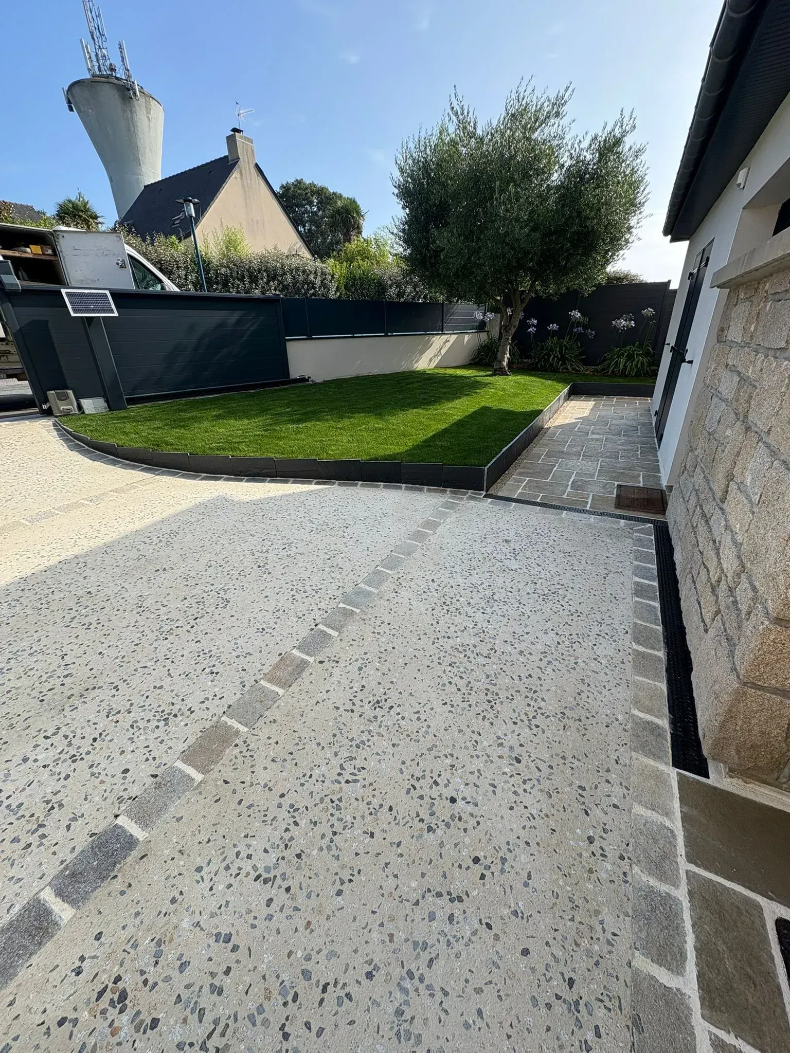 Allée de gravier longeant une maison et une pelouse, avec un chemin piétonnier en briques et des bâtiments au toit de chaume en arrière-plan.