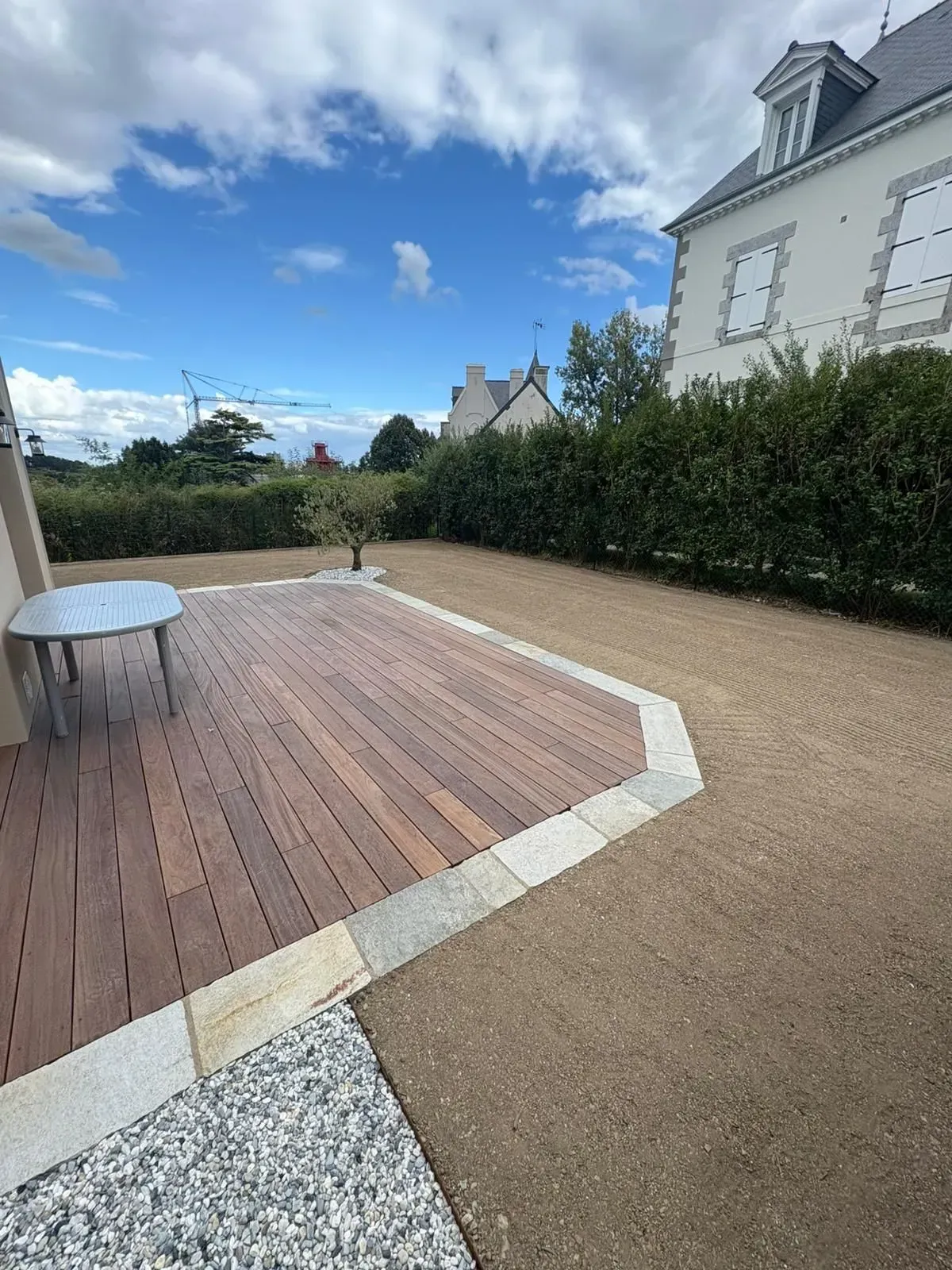 Terrasse en bois jouxtant un patio en gravier, bordé de haies et d'une maison blanche sous un ciel partiellement nuageux.