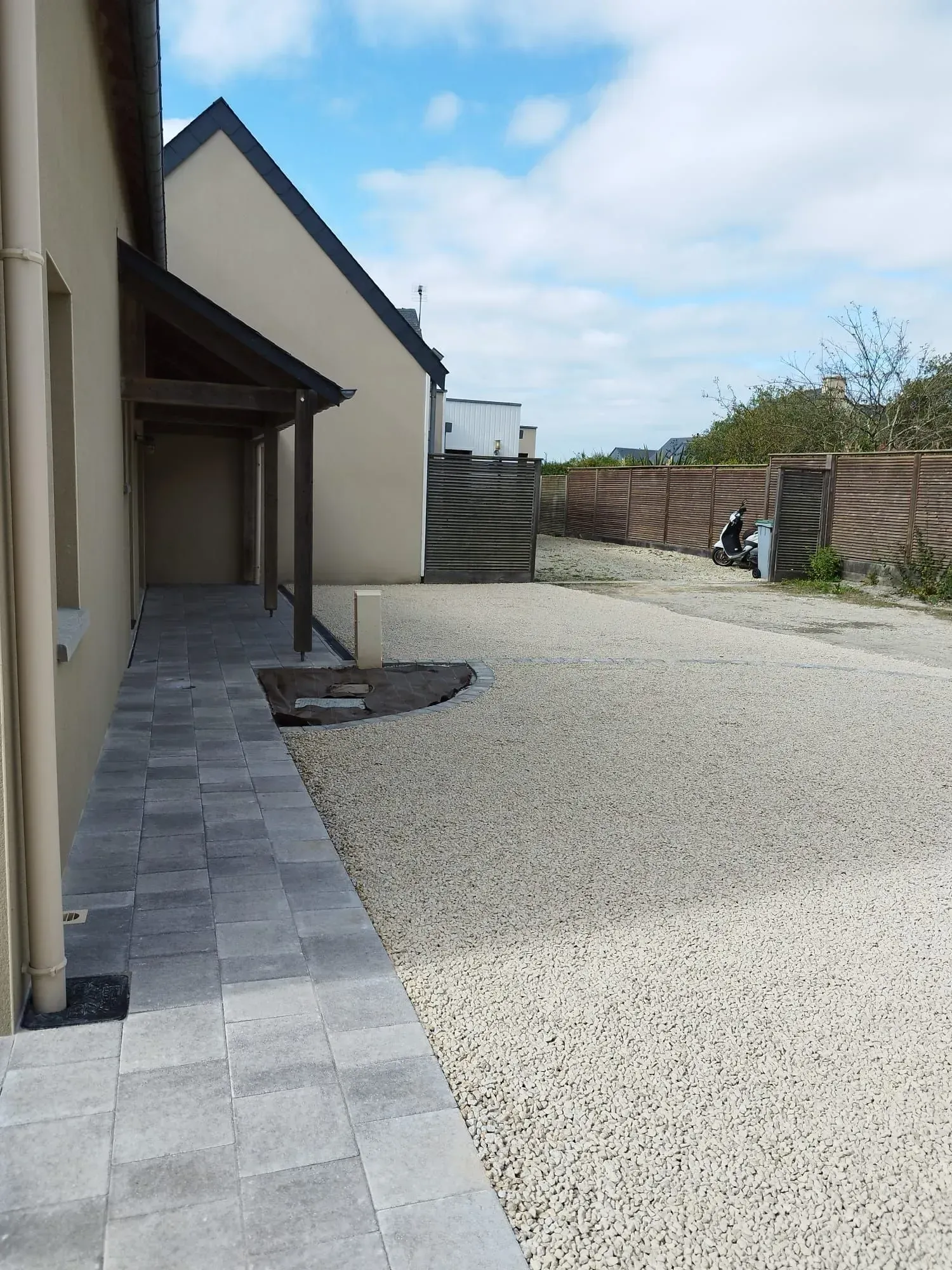 Allée de gravier à côté d'une maison aux pavés gris, aux murs couleur crème et à la cour clôturée, sous un ciel nuageux.