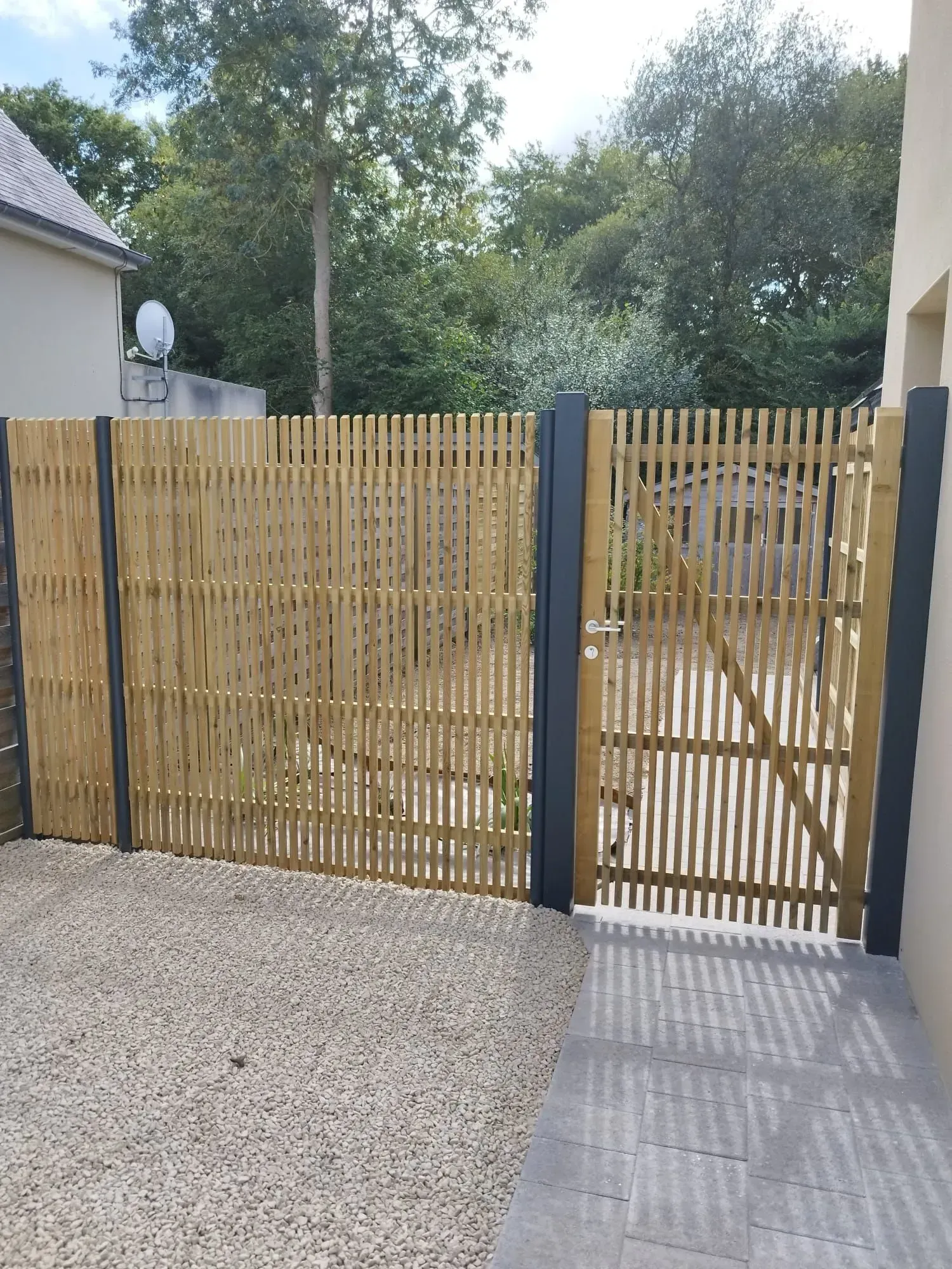 Clôture en bambou et portail assorti entre les maisons, donnant sur une allée de gravier et un chemin pavé.