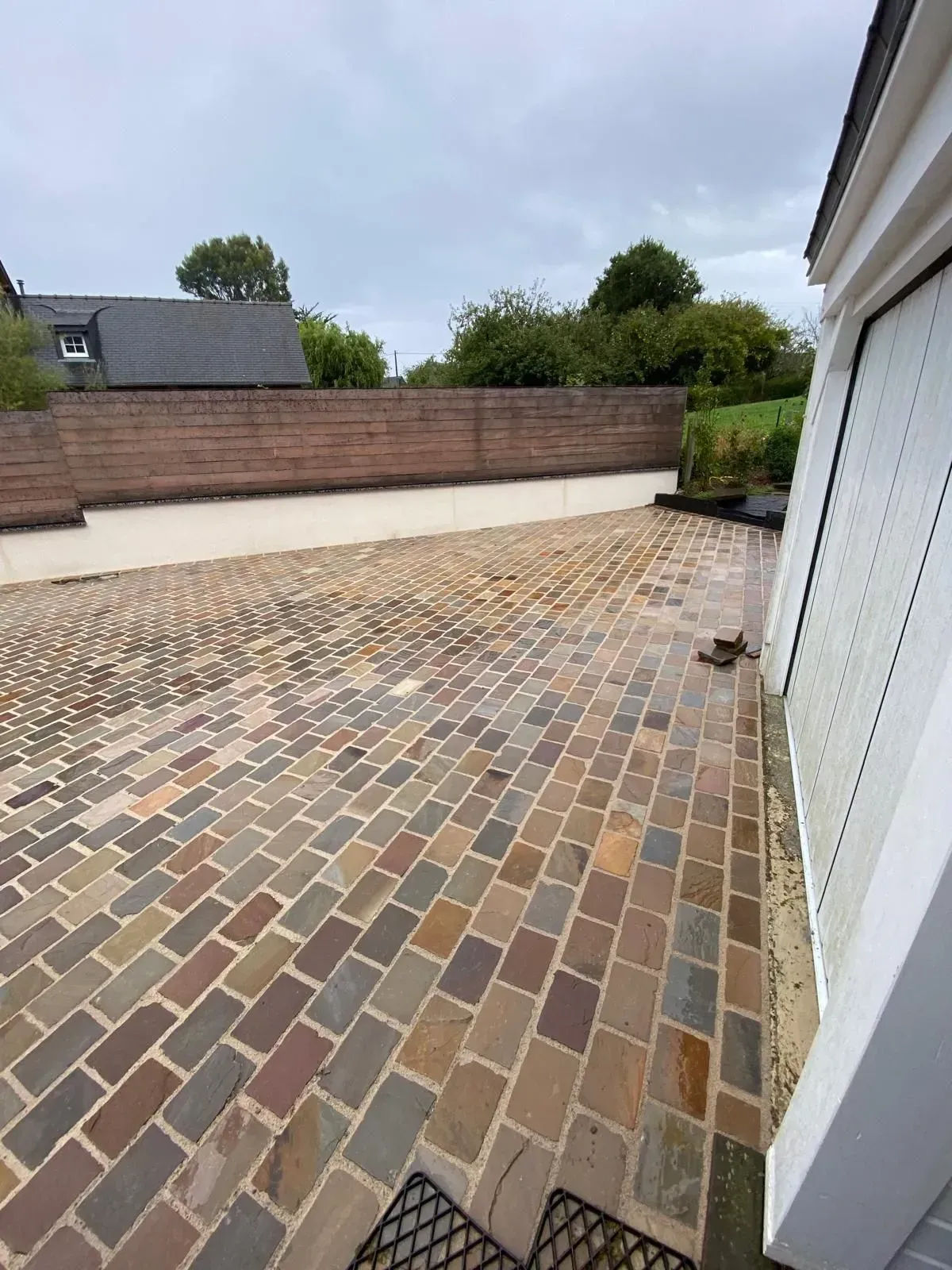 Terrasse pavée de briques multicolores jouxtant une maison blanche et un mur de jardin en briques.