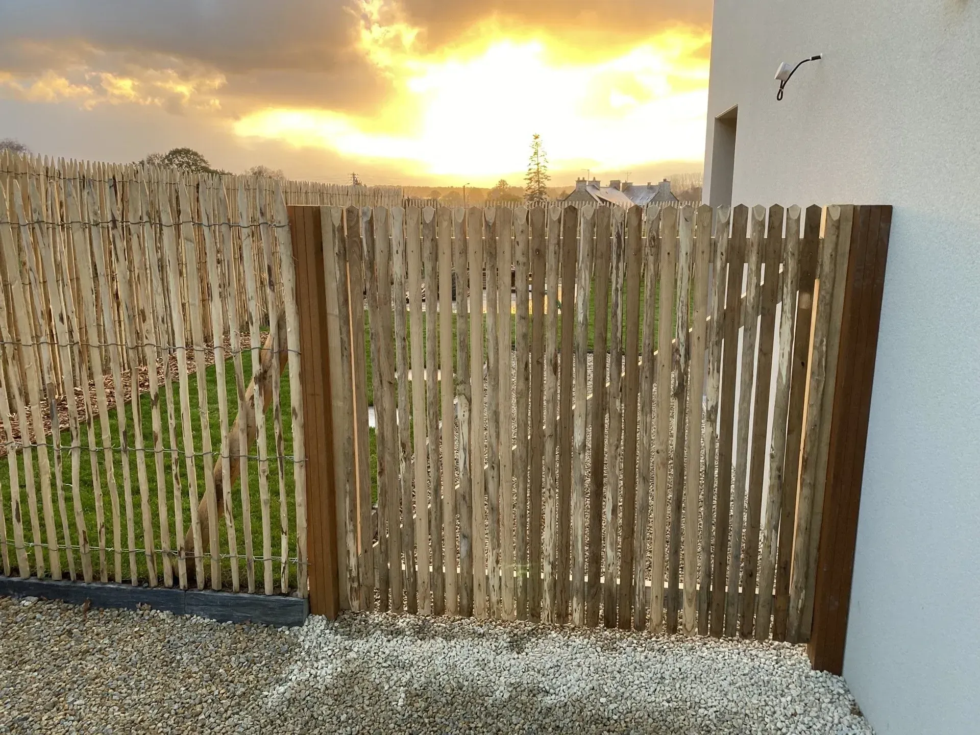 Clôture de jardin en bois au coucher du soleil, à côté d'un mur blanc et d'un chemin de gravier, avec de l'herbe verte derrière.