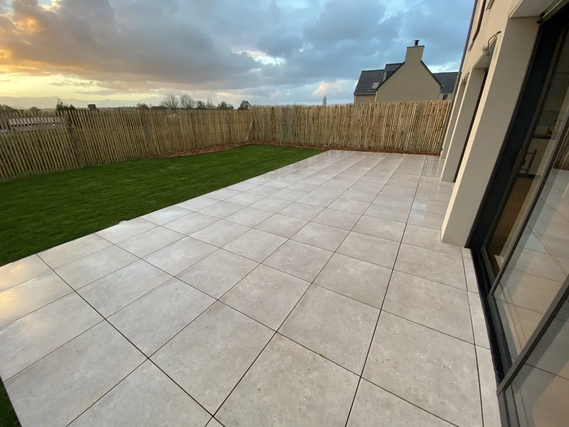 Terrasse arrière avec de grands carreaux gris clair, pelouse verte, clôture en bois et ciel nuageux au coucher du soleil.