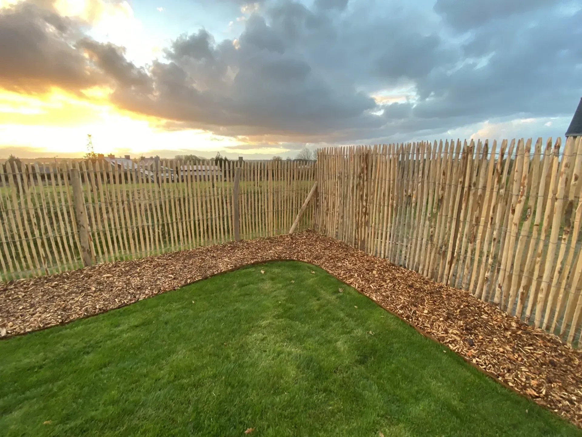 Jardin d'angle avec pelouse verte, clôture en bois et ciel de coucher de soleil spectaculaire aux nuages.