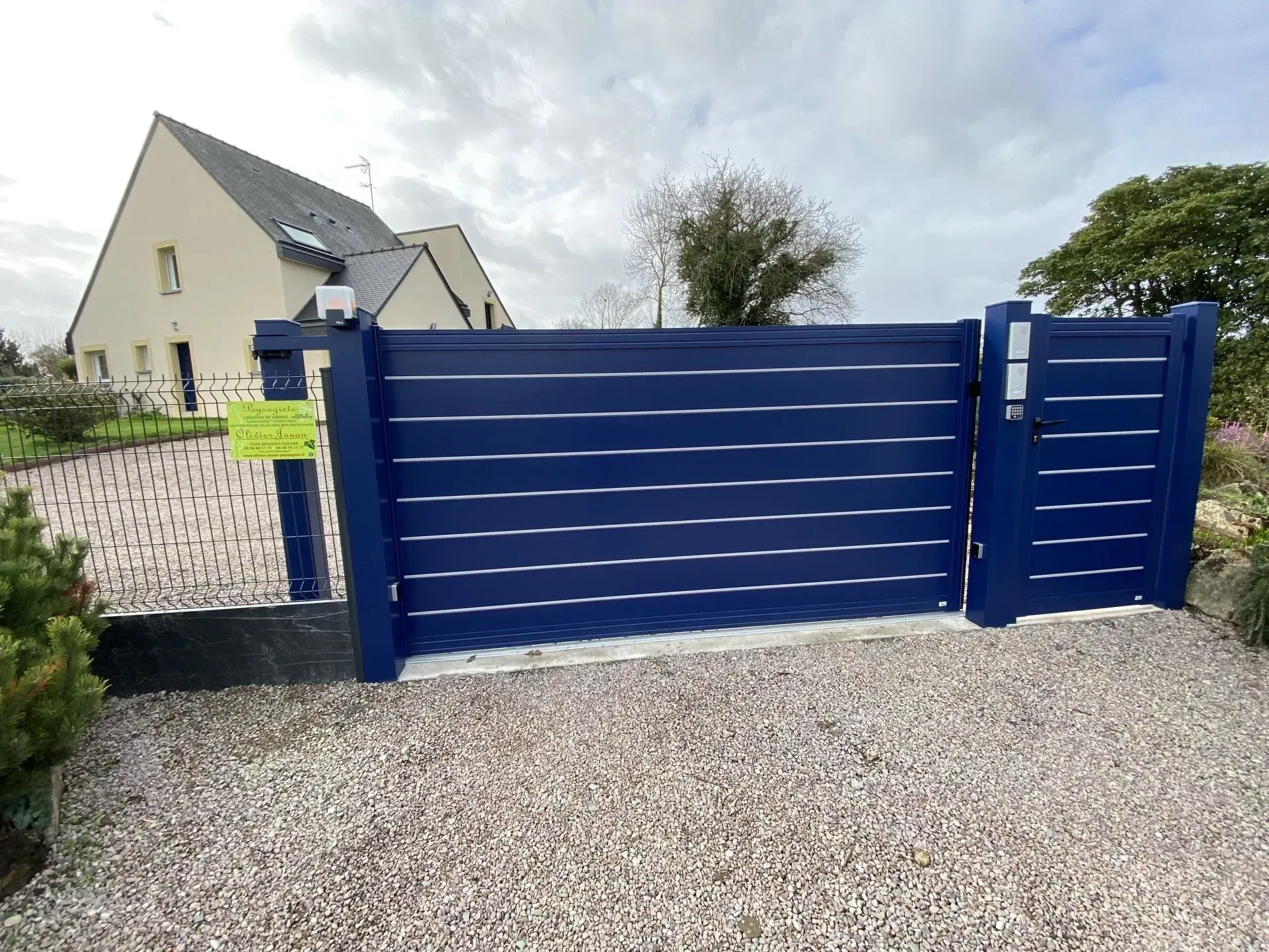 Portail coulissant bleu donnant sur une allée de gravier devant une maison et un jardin.
