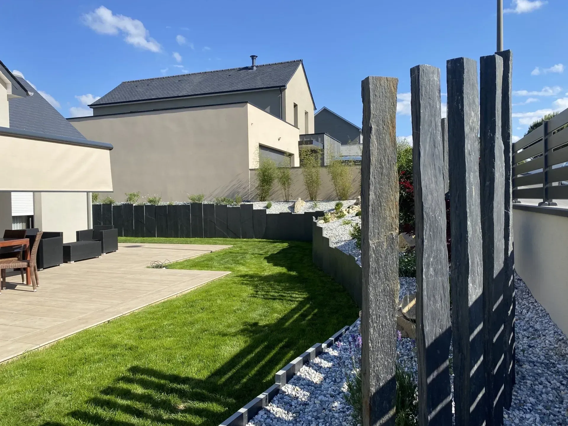 Terrasse arrière avec pelouse verte, maison beige et haute clôture sombre sous un ciel bleu clair.