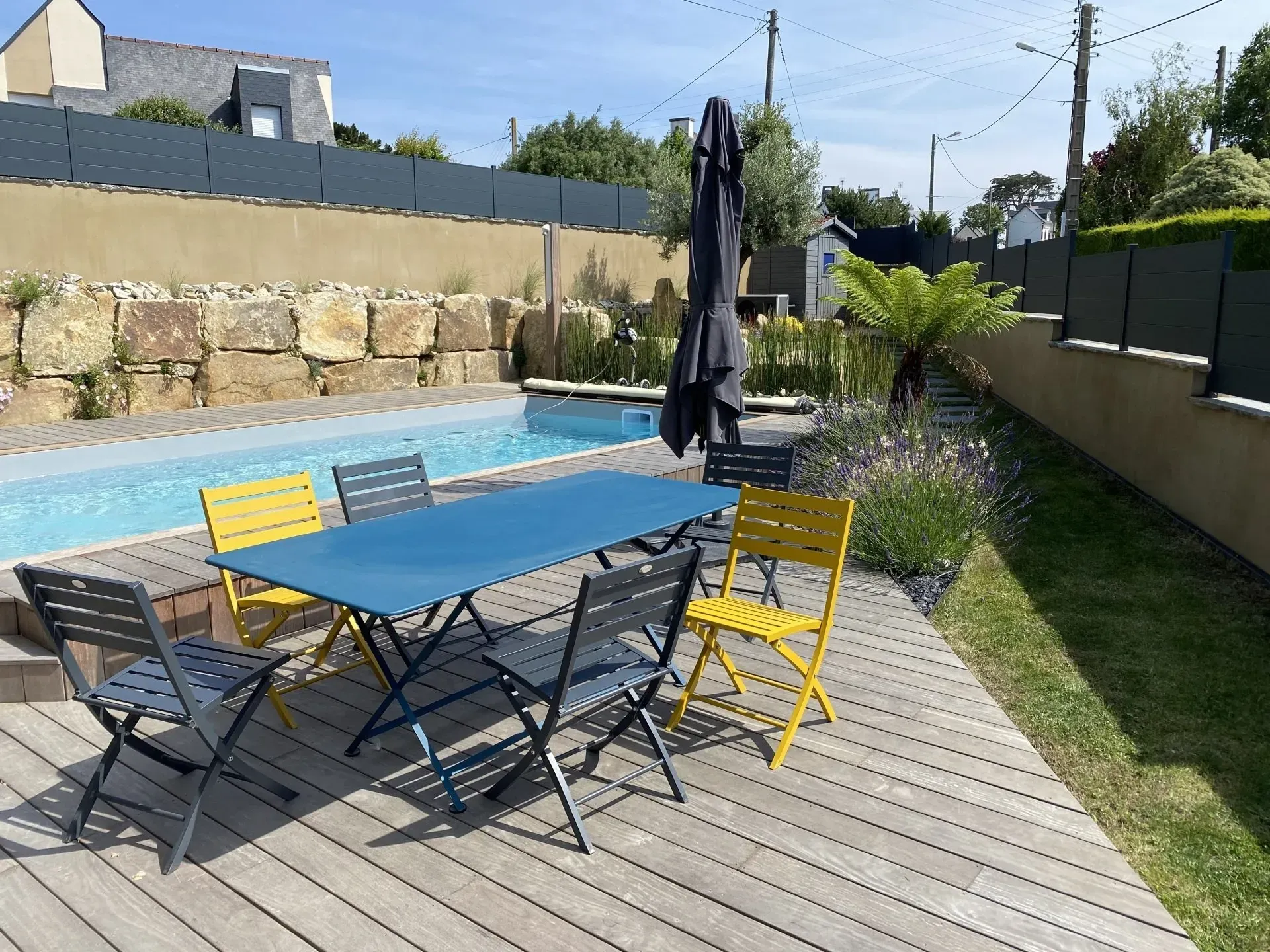 Terrasse extérieure avec table bleue, chaises colorées, parasol et piscine à côté d'un jardin gazonné.