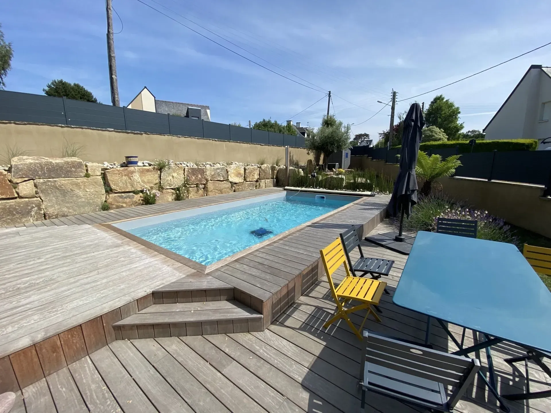 Piscine dans le jardin avec terrasse en bois, chaises jaunes, table bleue et muret en pierre au soleil.