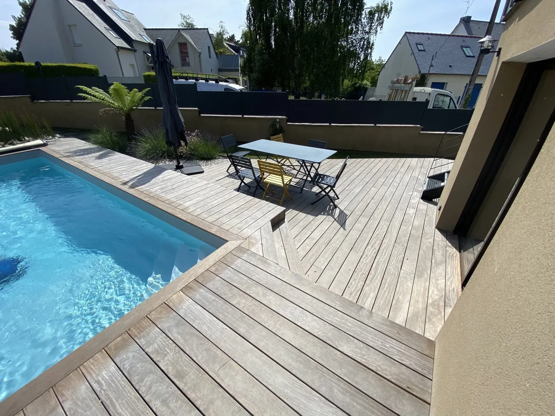 Terrasse de piscine dans le jardin avec table de patio, parasol et chaises longues baignées de soleil.