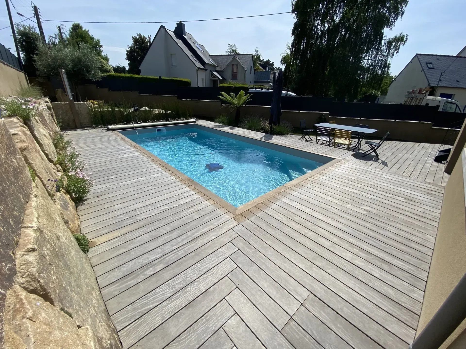 Piscine dans la cour arrière avec terrasse en pierre, chaises de patio et maisons en arrière-plan.
