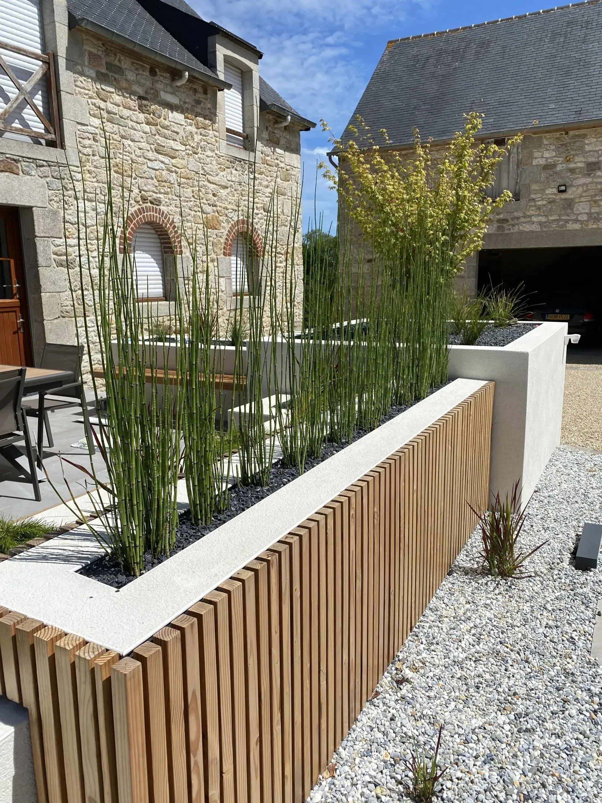Mur de jardin en pierre avec des lattes de bois, du gravier blanc et de hautes plantes vertes à côté d'une maison en pierre.