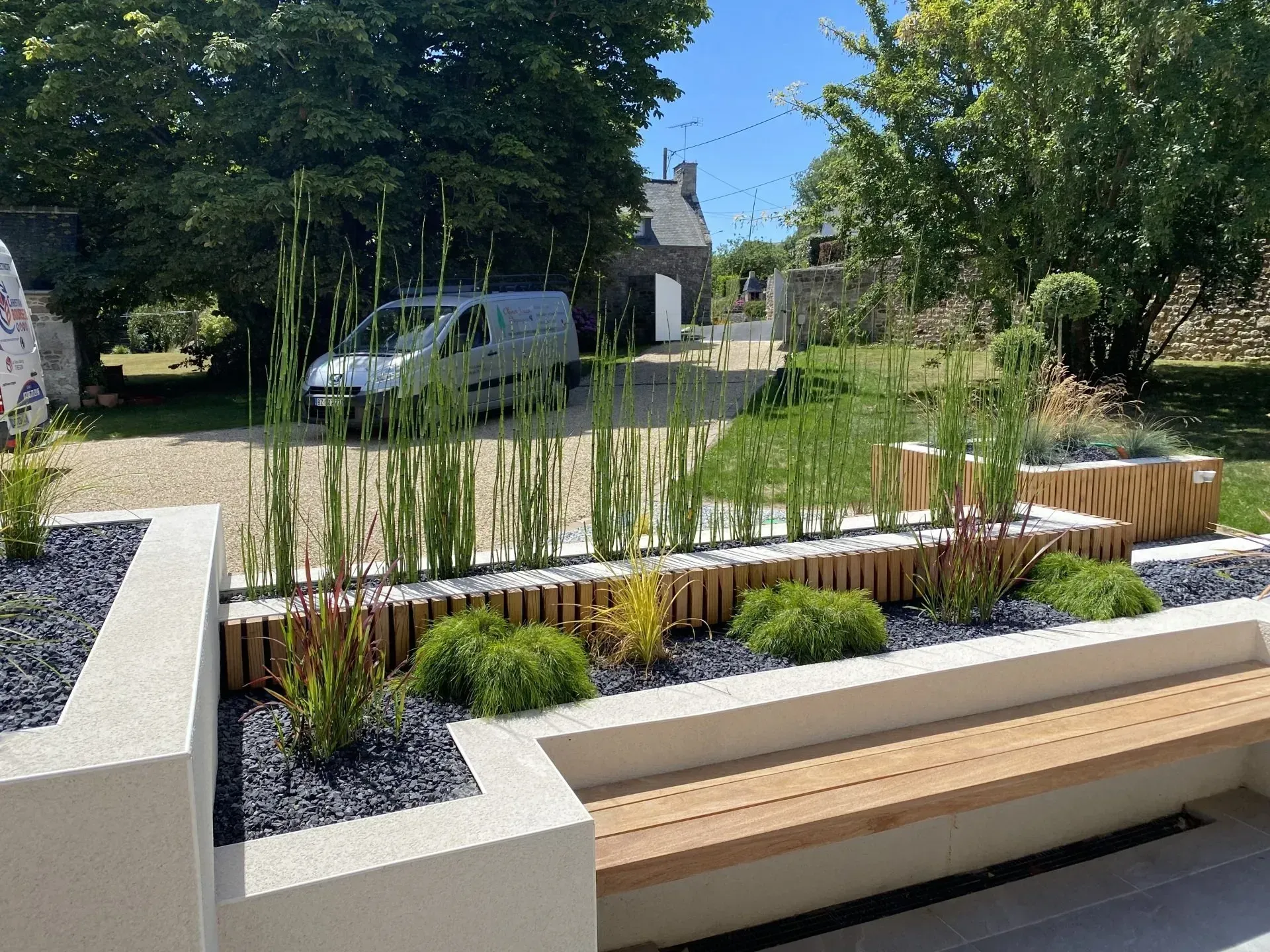 Jardin paysager moderne avec jardinières surélevées, hautes herbes, bancs et une camionnette en arrière-plan.