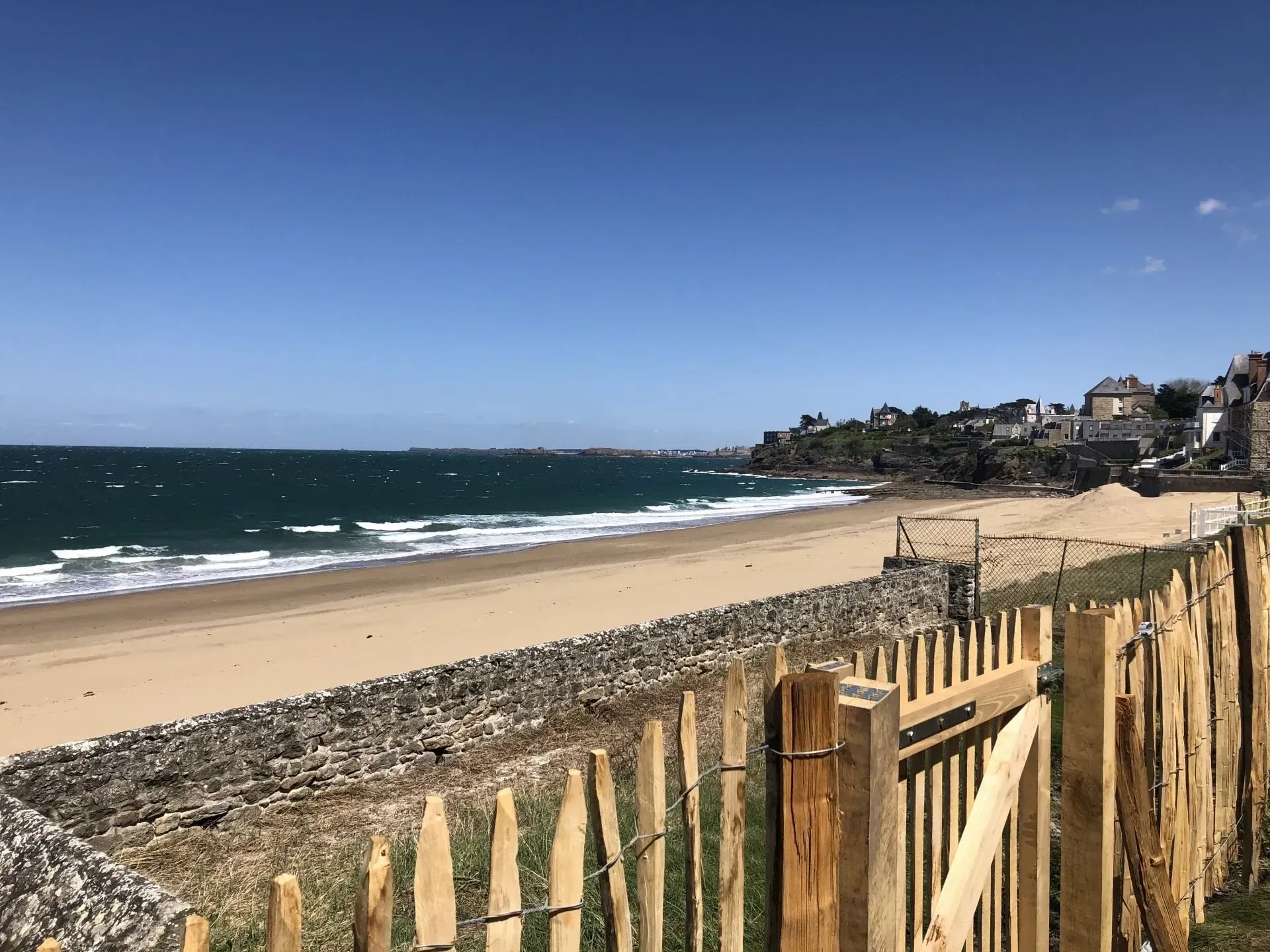 Plage ensoleillée avec des vagues, du sable et une clôture en bois le long du rivage sous un ciel bleu clair.