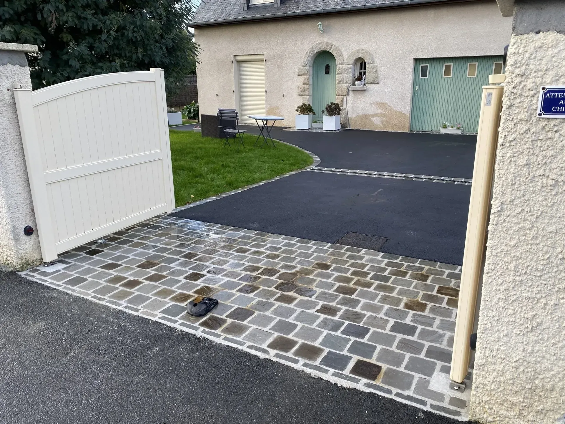 Un portail blanc ouvert donne sur une allée pavée et une maison beige aux volets verts.