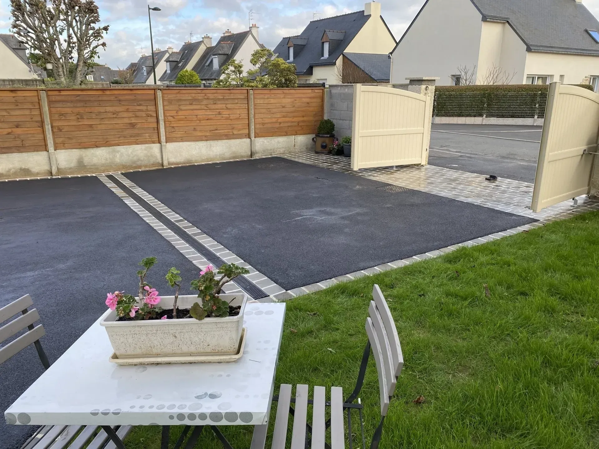 Terrasse avec table et chaises d'extérieur, fleurs en pot, portail ouvert et allée pavée donnant sur une pelouse.