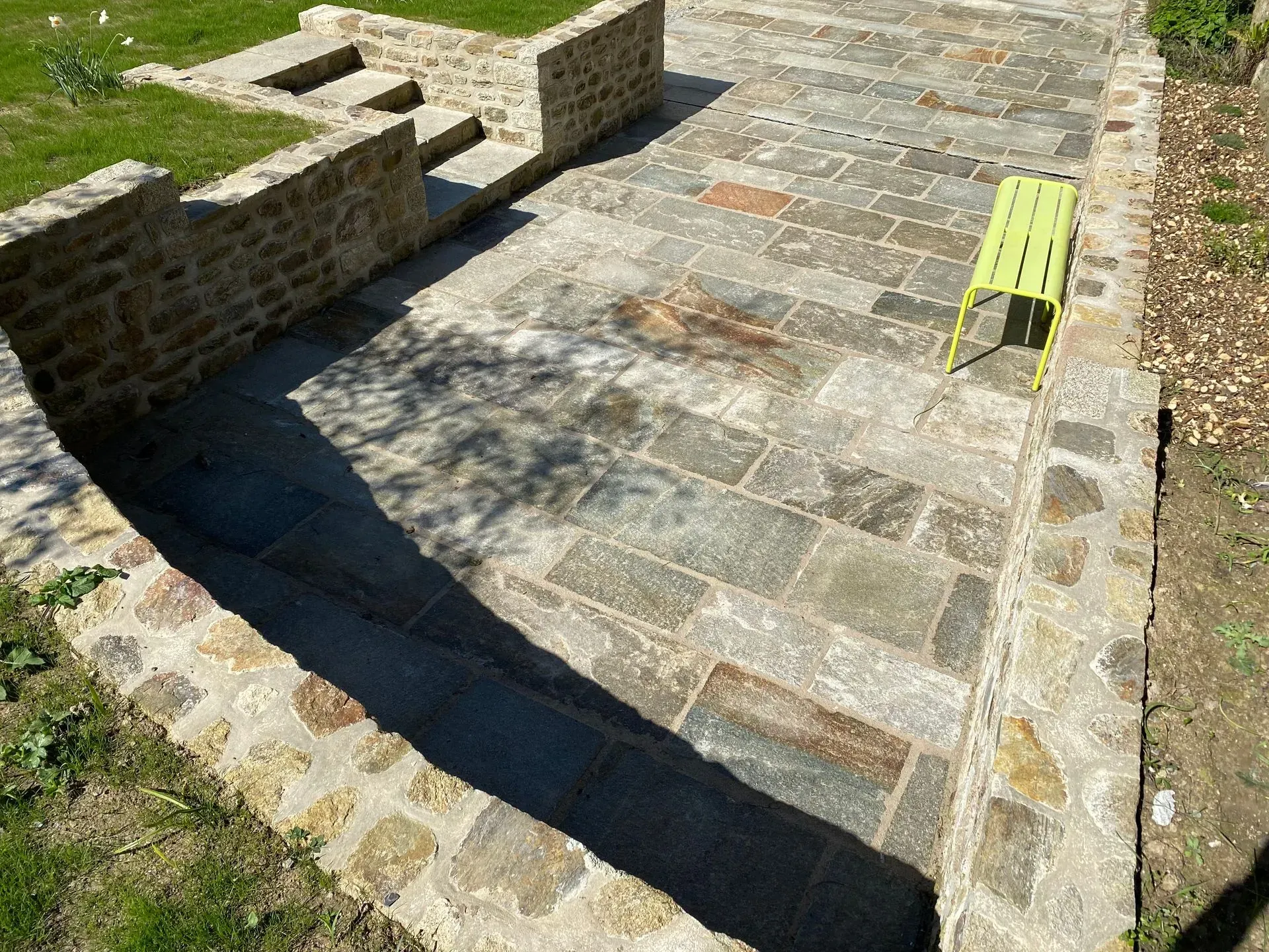 Terrasse en pierre avec des marches longeant un mur de briques, et un banc vert citron baigné de soleil et d'ombre.
