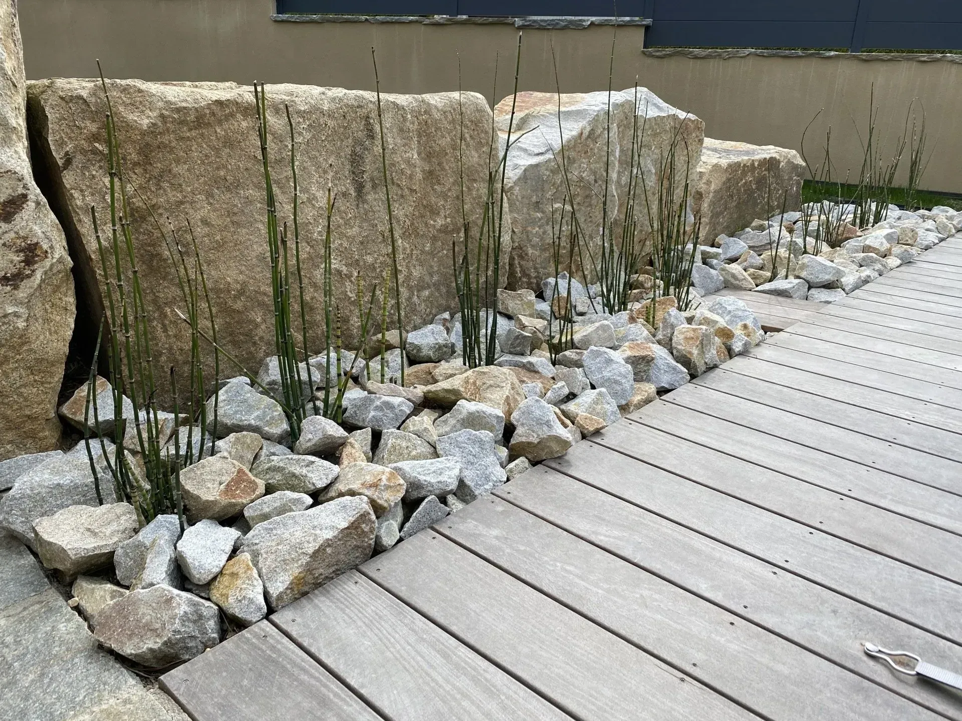 Jardin de rocaille avec de gros rochers et de hautes herbes, bordé d'une terrasse en bois et d'une bordure en pierre.