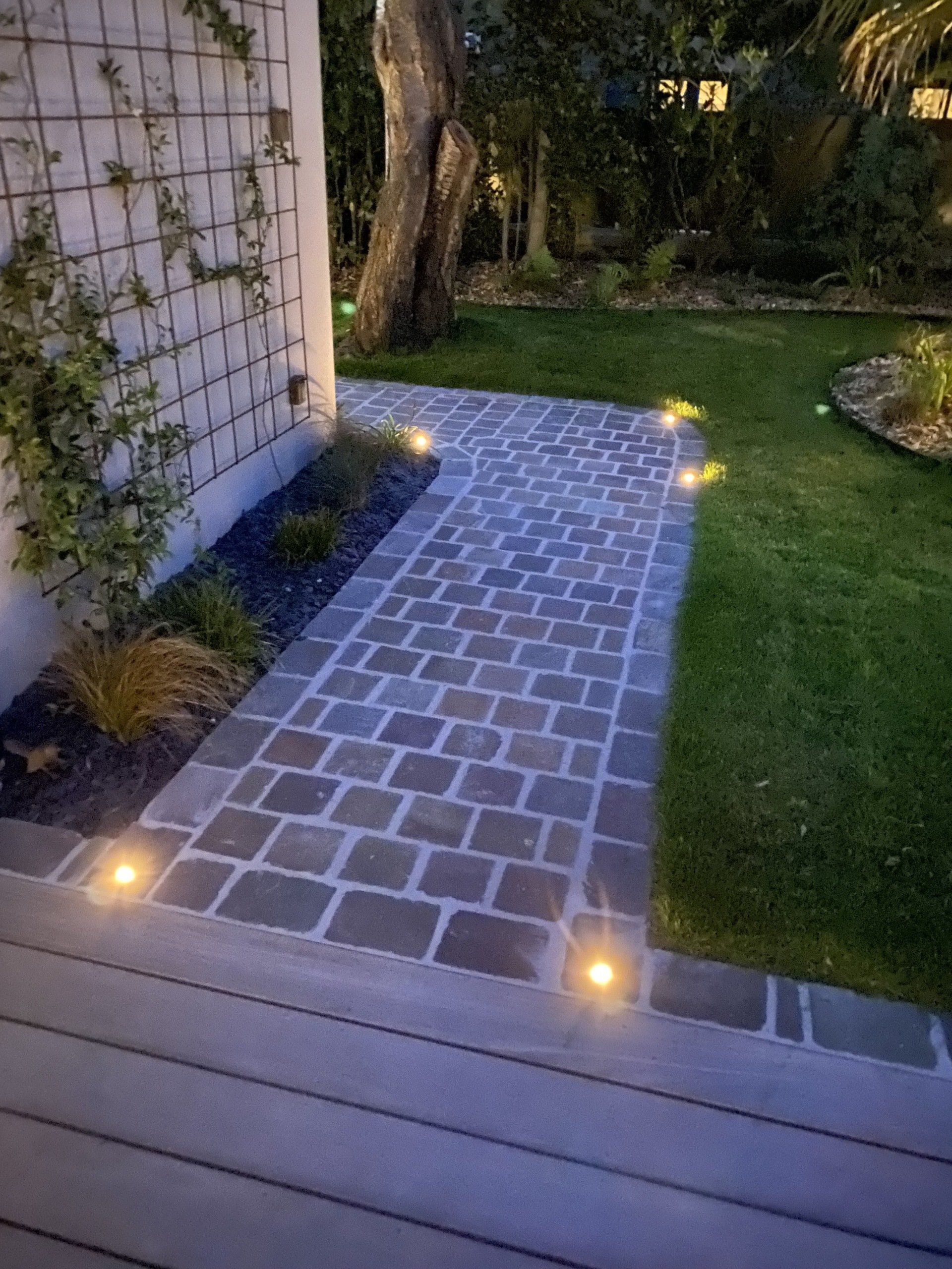 Allée de terrasse en pierre éclairée à côté d'une maison, menant à une cour arrière gazonnée au crépuscule.